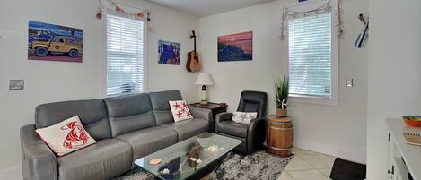 A living room with a grey sofa, a glass coffee table, a black armchair, a wooden barrel, and a ceiling fan.