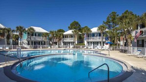 A pool area with a clear blue pool, surrounded by two-story buildings with balconies, palm trees, and American flags. - Bungalows at Seagrove by Southern Vacation Rentals (Santa Rosa Beach)