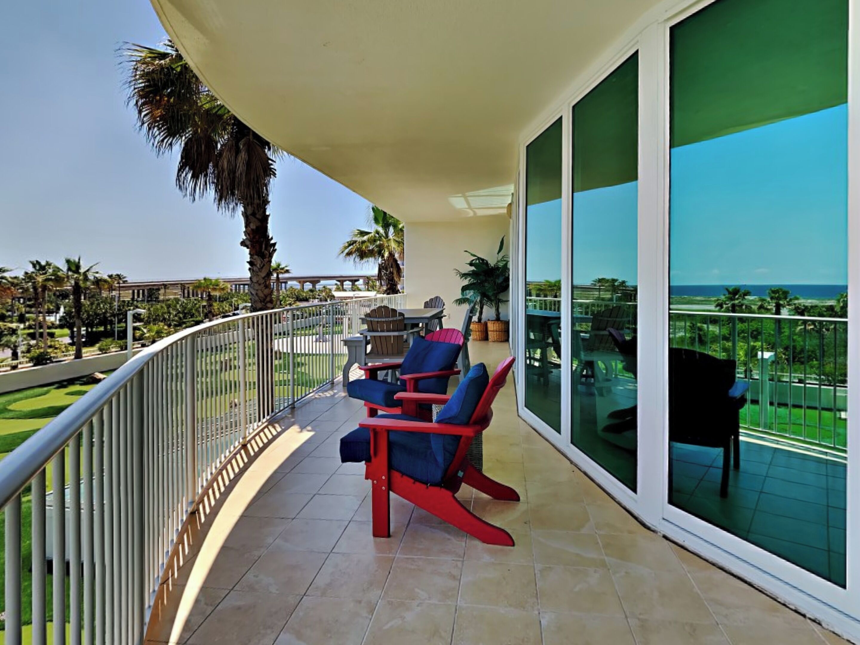 A balcony with red chairs, a table, and a view of the ocean and greenery.
