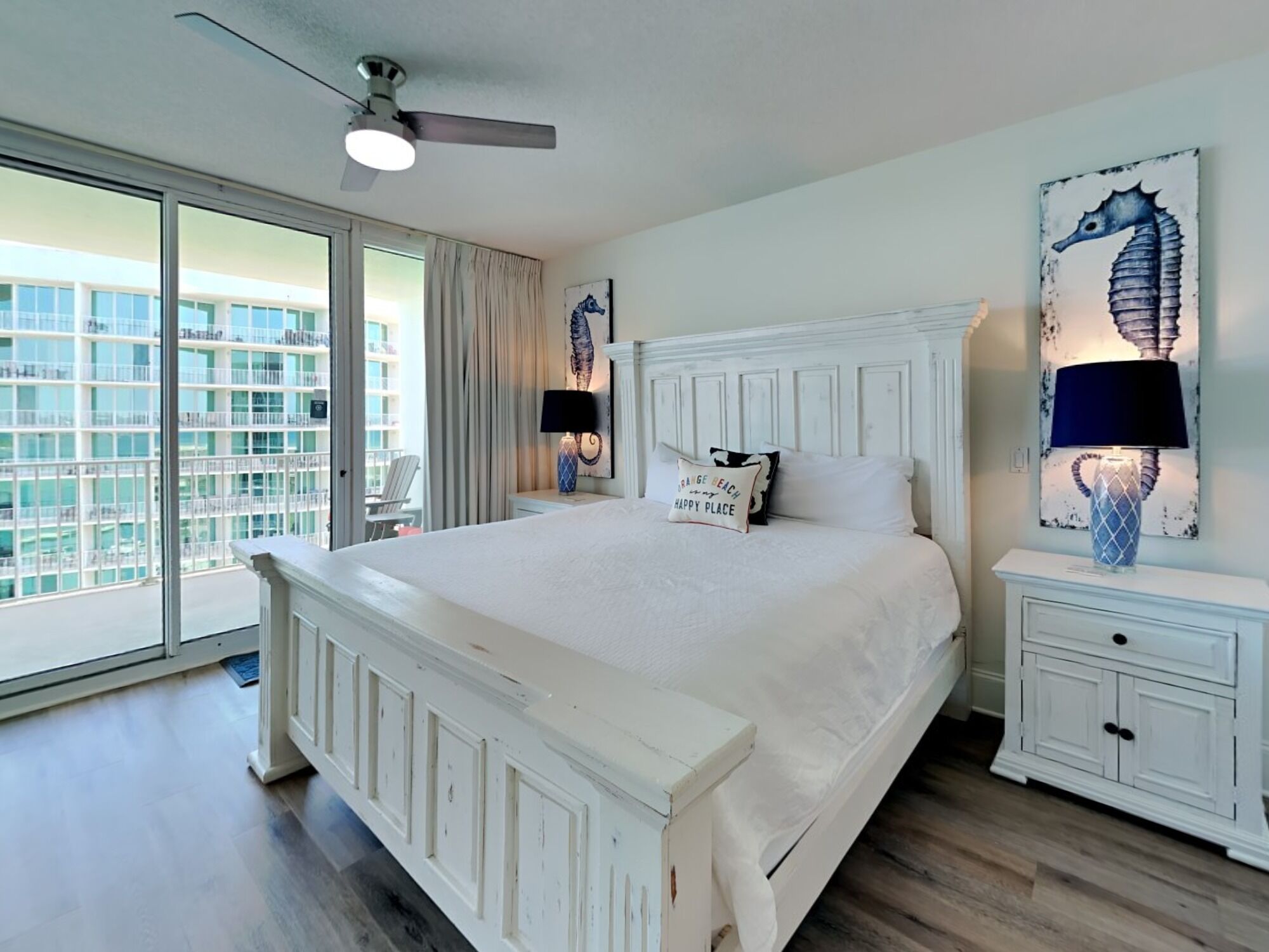 A spacious bedroom with a large bed, a ceiling fan, and a view of a building through a sliding glass door.