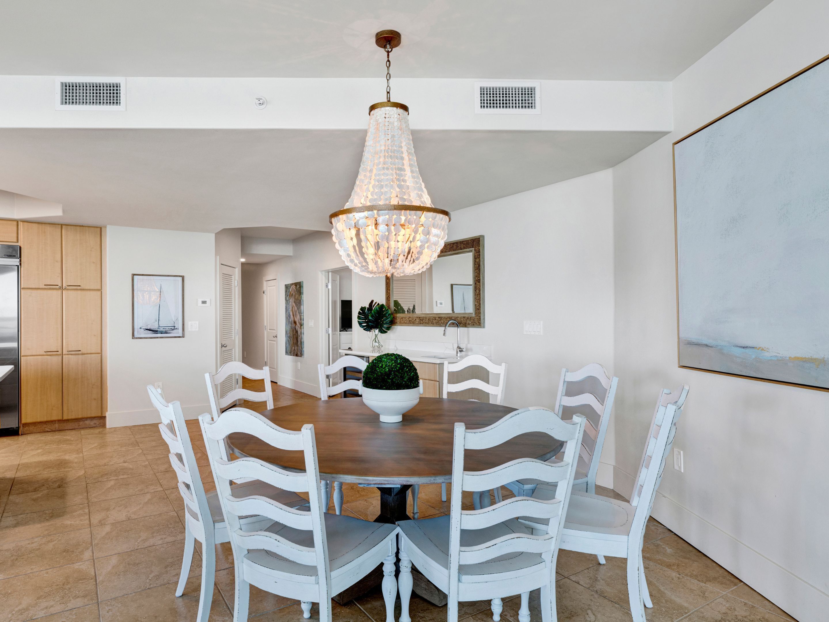A dining area with a round table, white chairs, a chandelier, a potted plant, and a large painting on the wall.