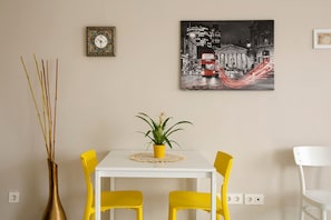 Un comedor con una mesa blanca, sillas amarillas, una planta en maceta, un reloj de pared y un cuadro con un paisaje urbano.