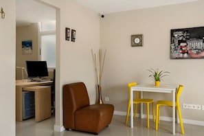 Una sala de estar moderna con un sofá marrón, una mesa de comedor con sillas amarillas, un escritorio blanco con una computadora y un florero con ramas.