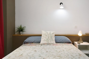 Una cama con ropa de cama floral, una mesita de noche con lámpara, una planta en maceta y un aplique de pared.