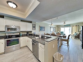 A spacious kitchen with white cabinets, a granite countertop, stainless steel appliances, and a dining area with a view of the beach.
