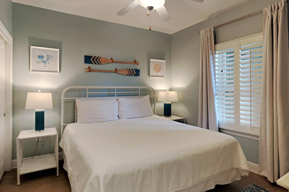 A neatly arranged bedroom with a bed, two bedside tables with lamps, a ceiling fan, and a window with blinds.