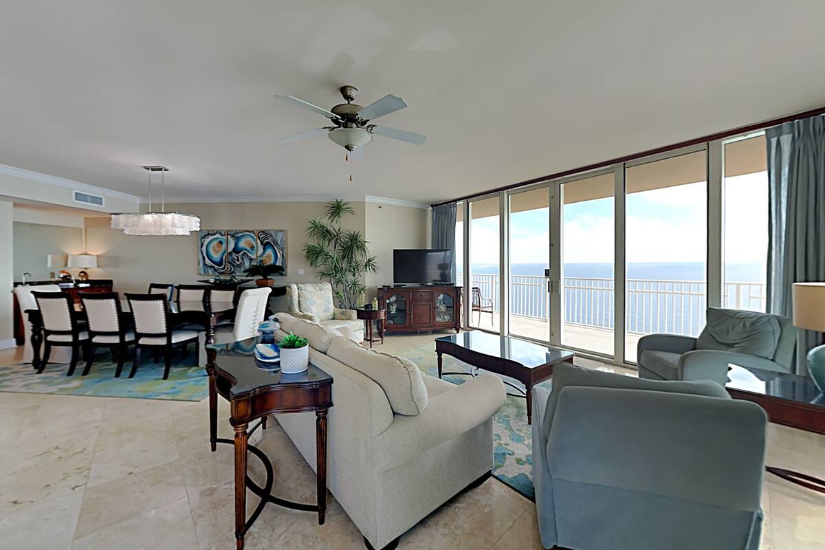 A spacious living room with a dining area, a ceiling fan, a large window with a view of the ocean, and a balcony.