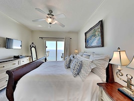 A hotel room with a bed, a television, a ceiling fan, a painting, and a view of the ocean through the balcony doors.
