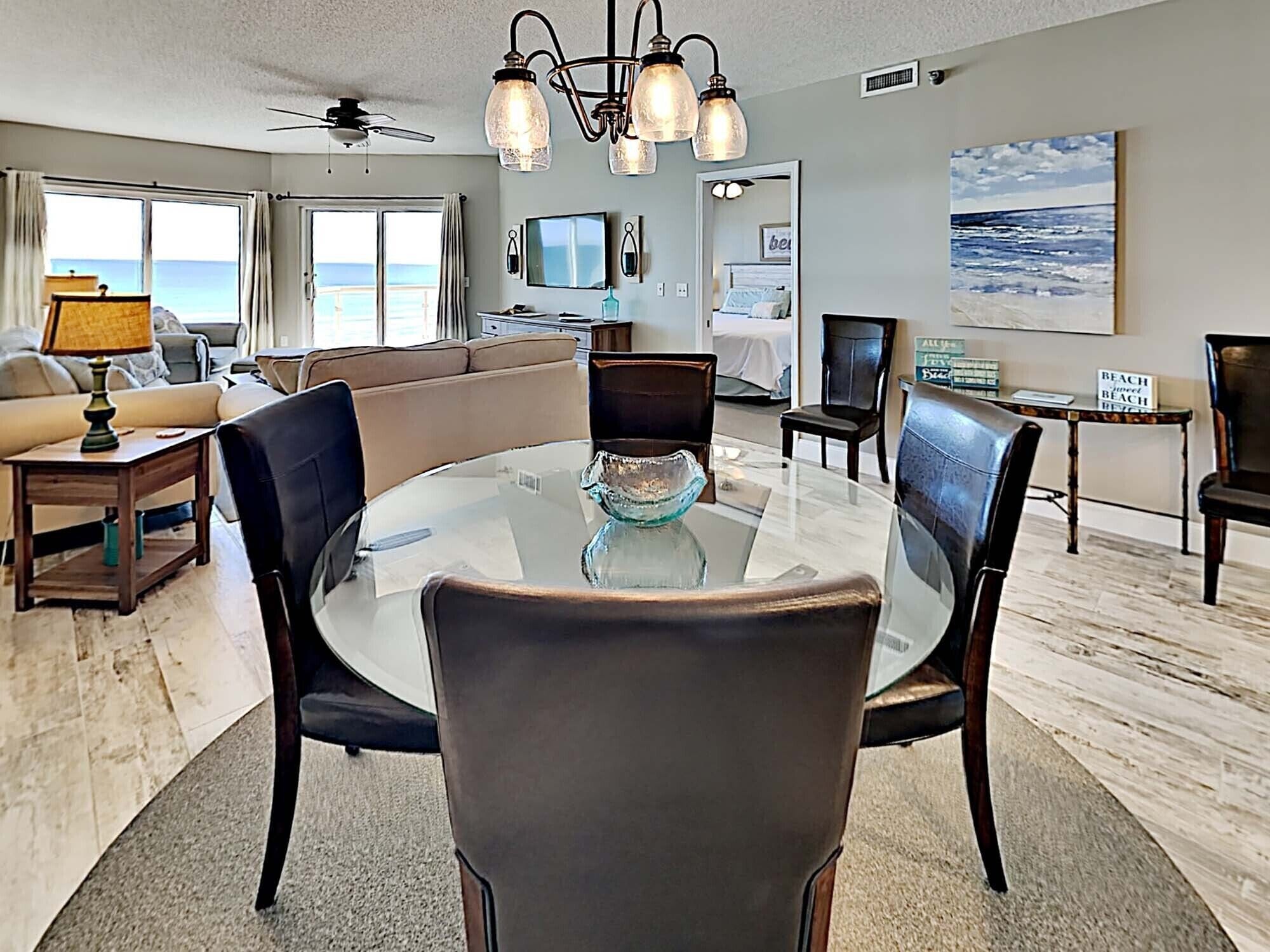 A spacious living room with a glass-top dining table, leather chairs, a ceiling fan, and a view of the ocean through large windows.