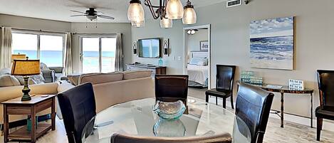 A spacious living room with a glass-top dining table, leather chairs, a ceiling fan, and a view of the ocean through large windows.