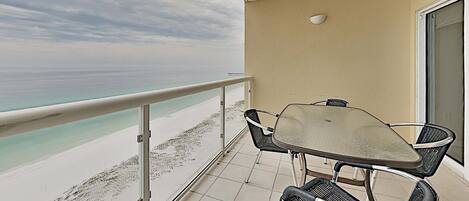 Un balcón con mesa y sillas de vidrio, con vista a una playa de aguas turquesas cristalinas y cielo nublado.