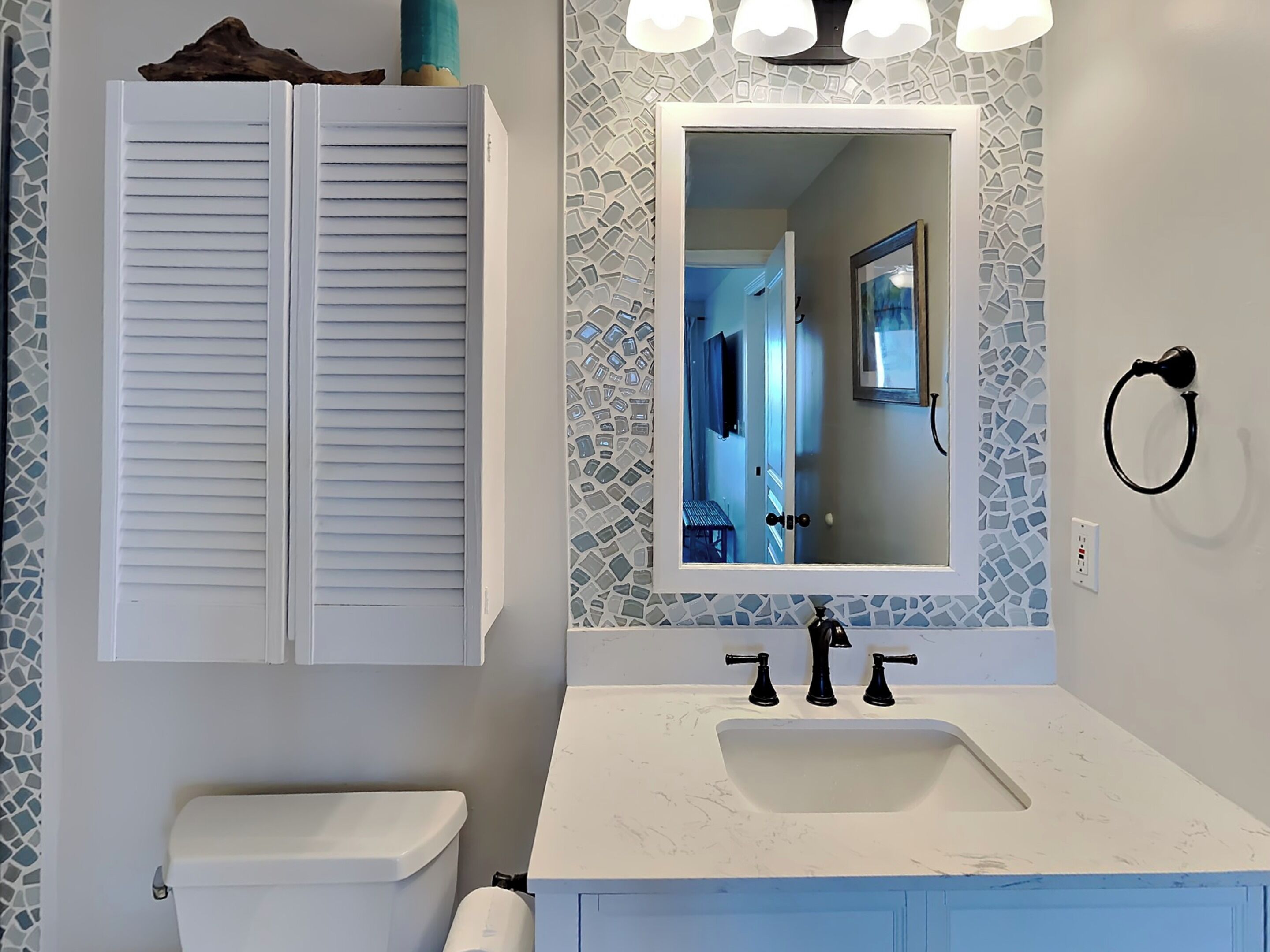A bathroom with a white toilet, a white sink with a marble countertop, a mirror with a mosaic backsplash, and a towel ring on the wall.