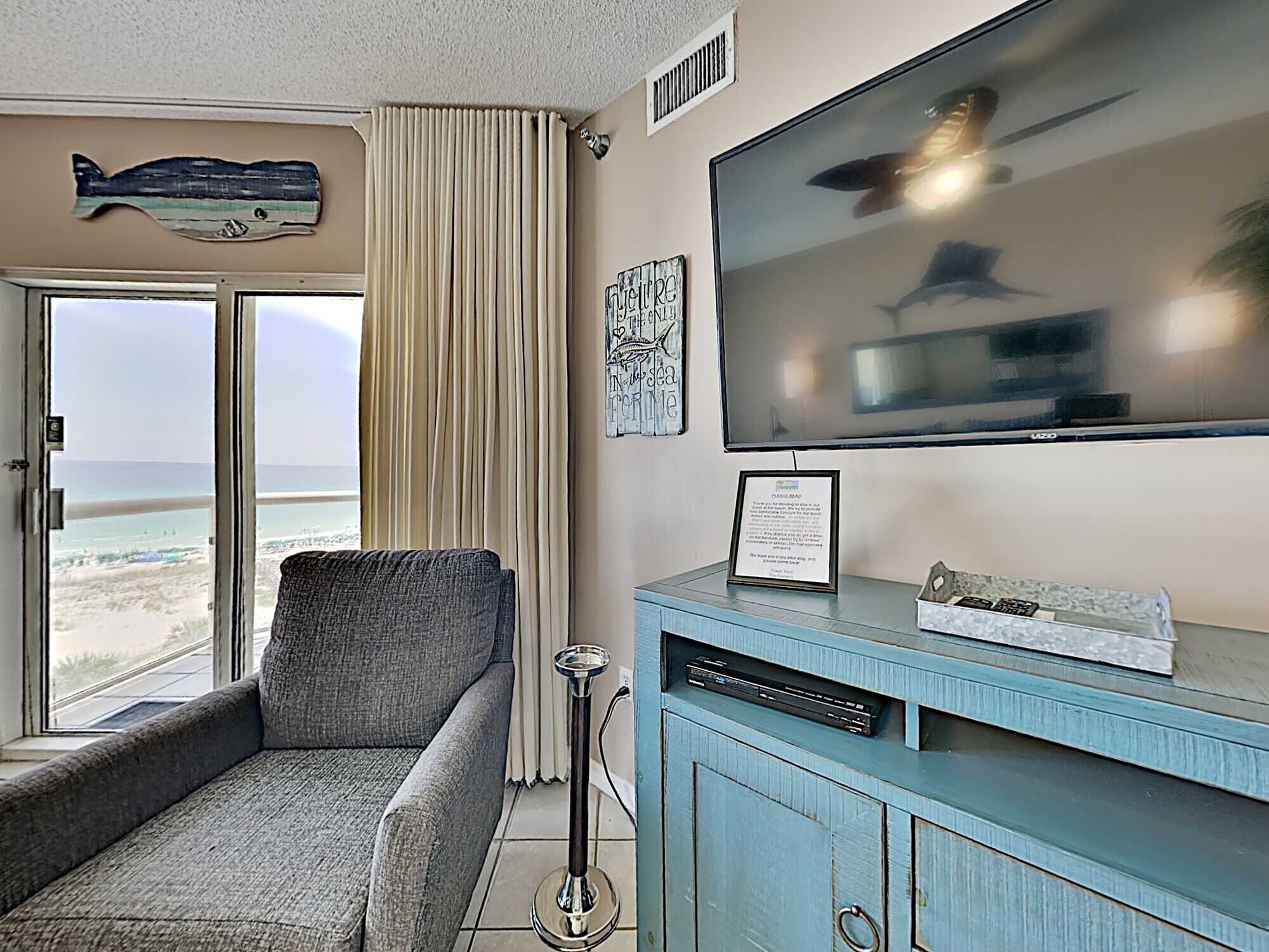 Habitación con televisor grande, sofá, mueble con control remoto y vista a la playa.
