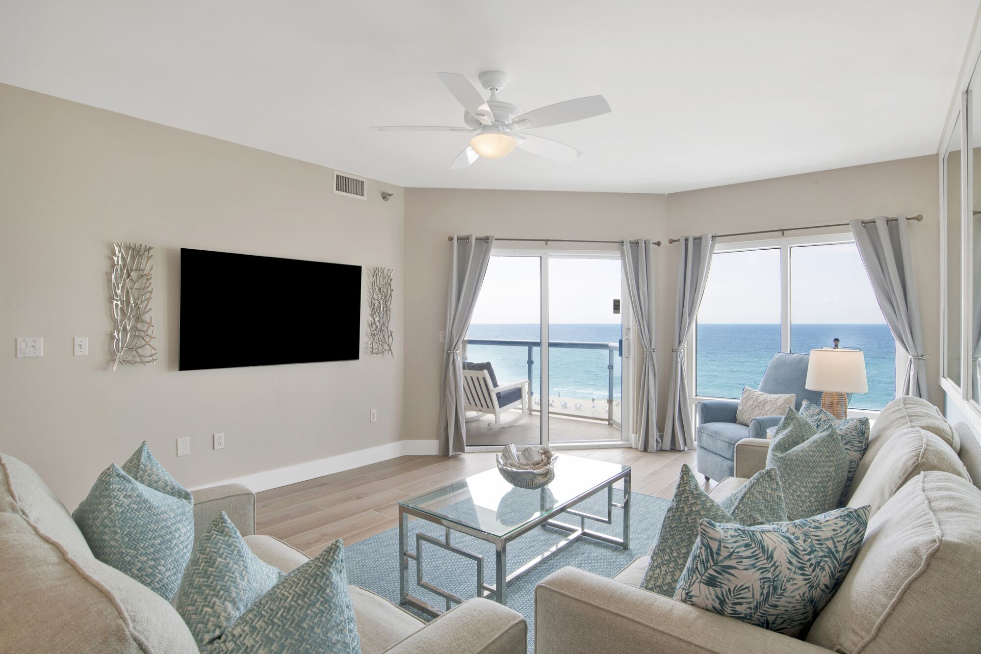 A spacious living room with a large flat-screen TV, a ceiling fan, and a view of the ocean through large sliding doors.