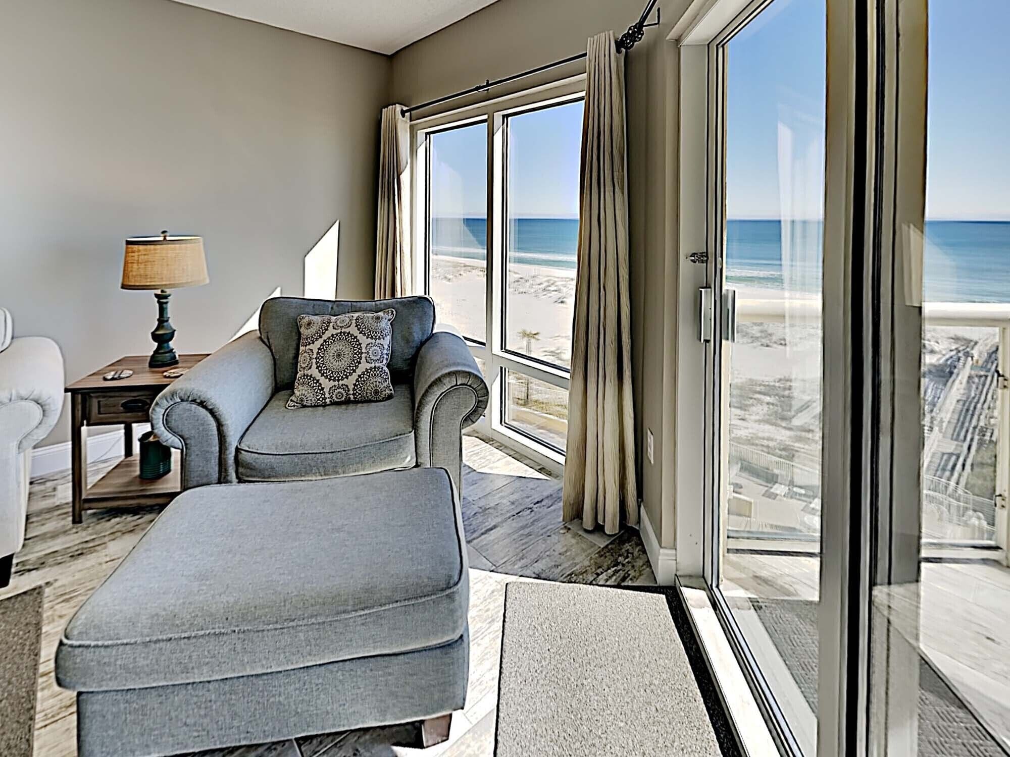 A room with a view of the beach, a grey sofa with a patterned pillow, a small table with a lamp, and a large window.