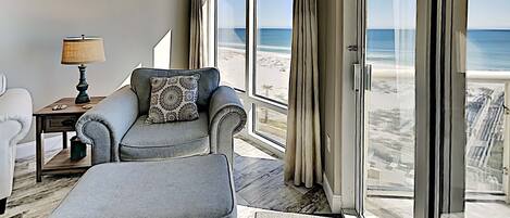 A room with a view of the beach, a grey sofa with a patterned pillow, a small table with a lamp, and a large window.