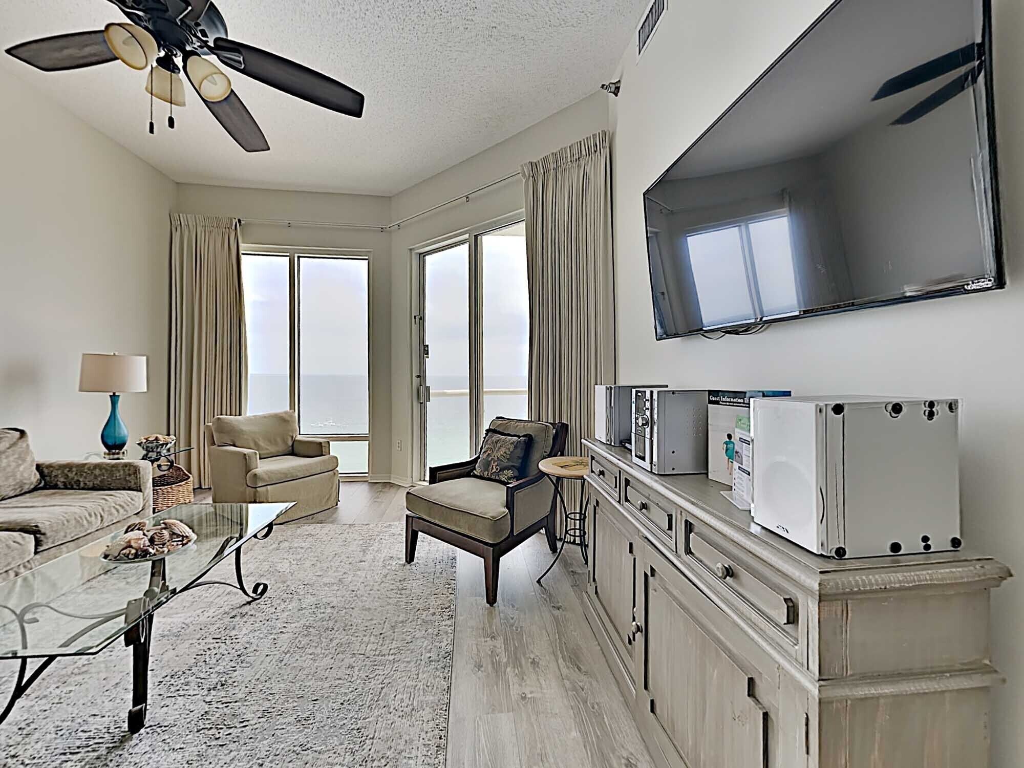 A spacious living room with a ceiling fan, a flat-screen TV, and a large window offering a view of the ocean.