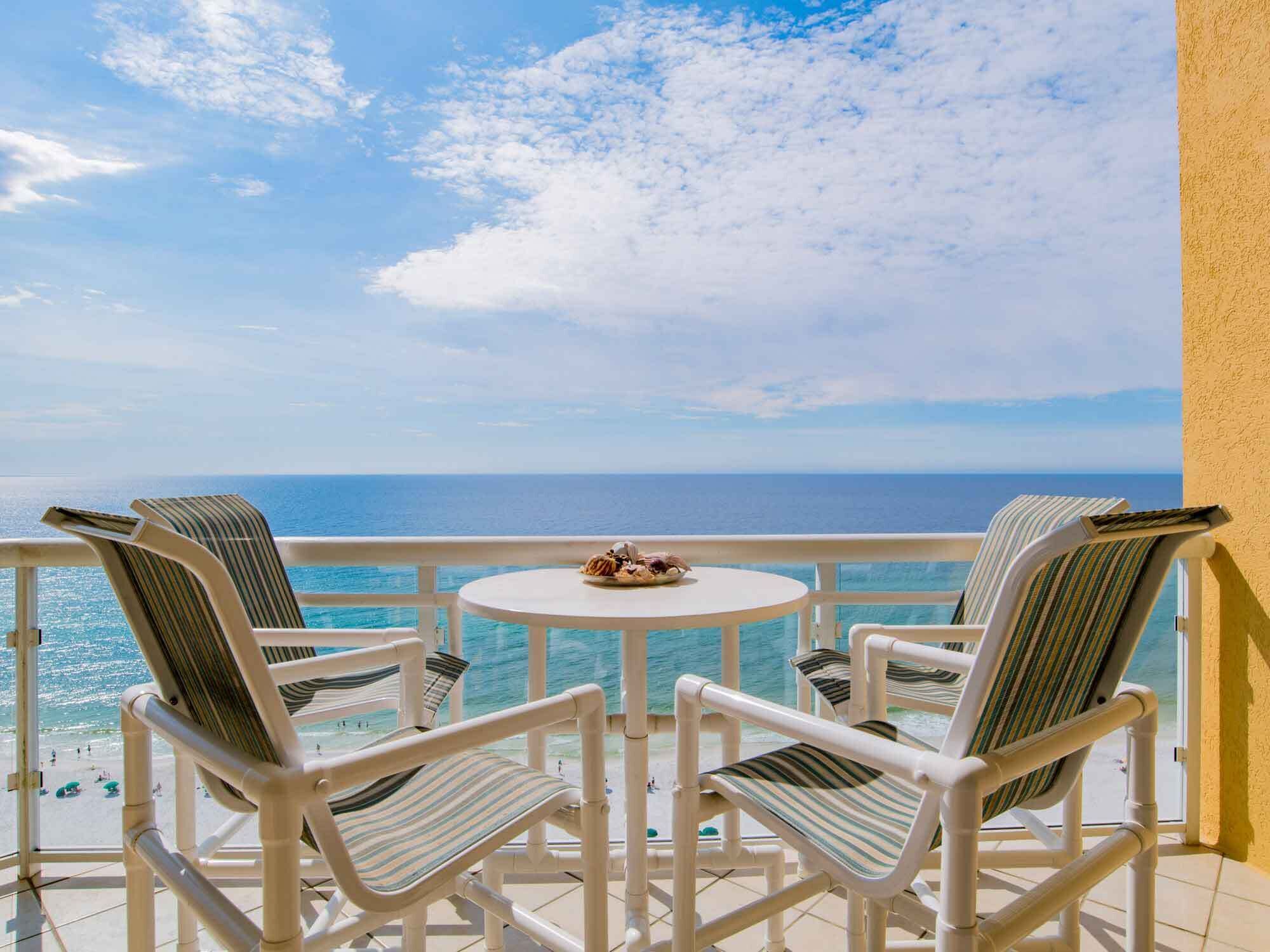 A balcony with two striped chairs and a small round table, overlooking a beach and the sea.
