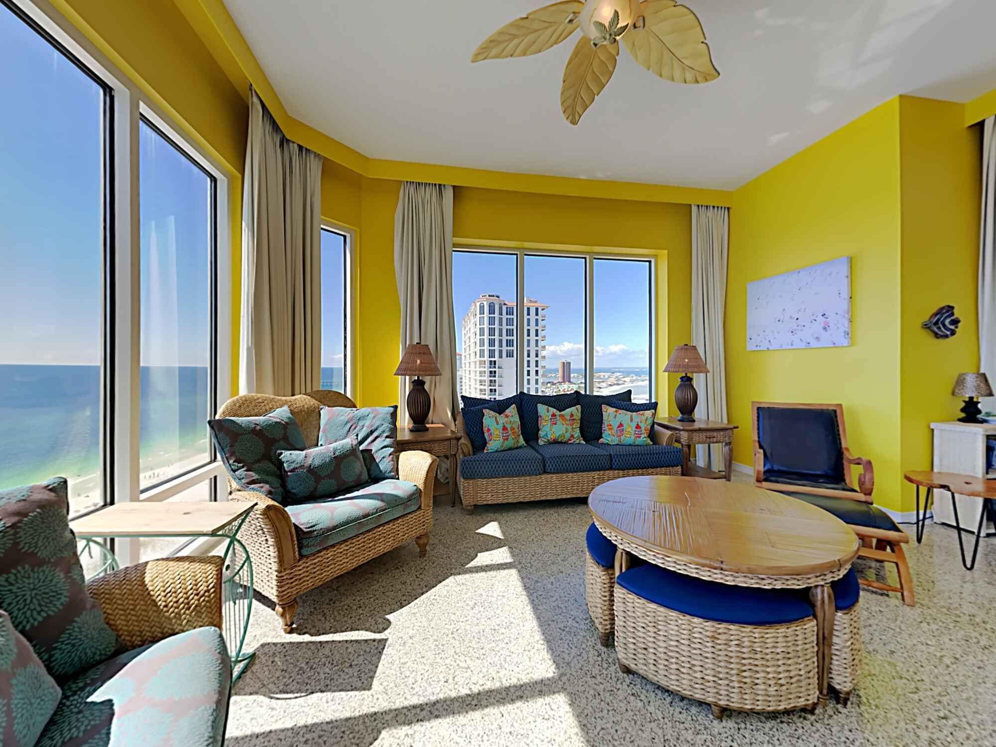 Una habitación luminosa de color amarillo con amplias ventanas, mobiliario de mimbre y un ventilador de techo.