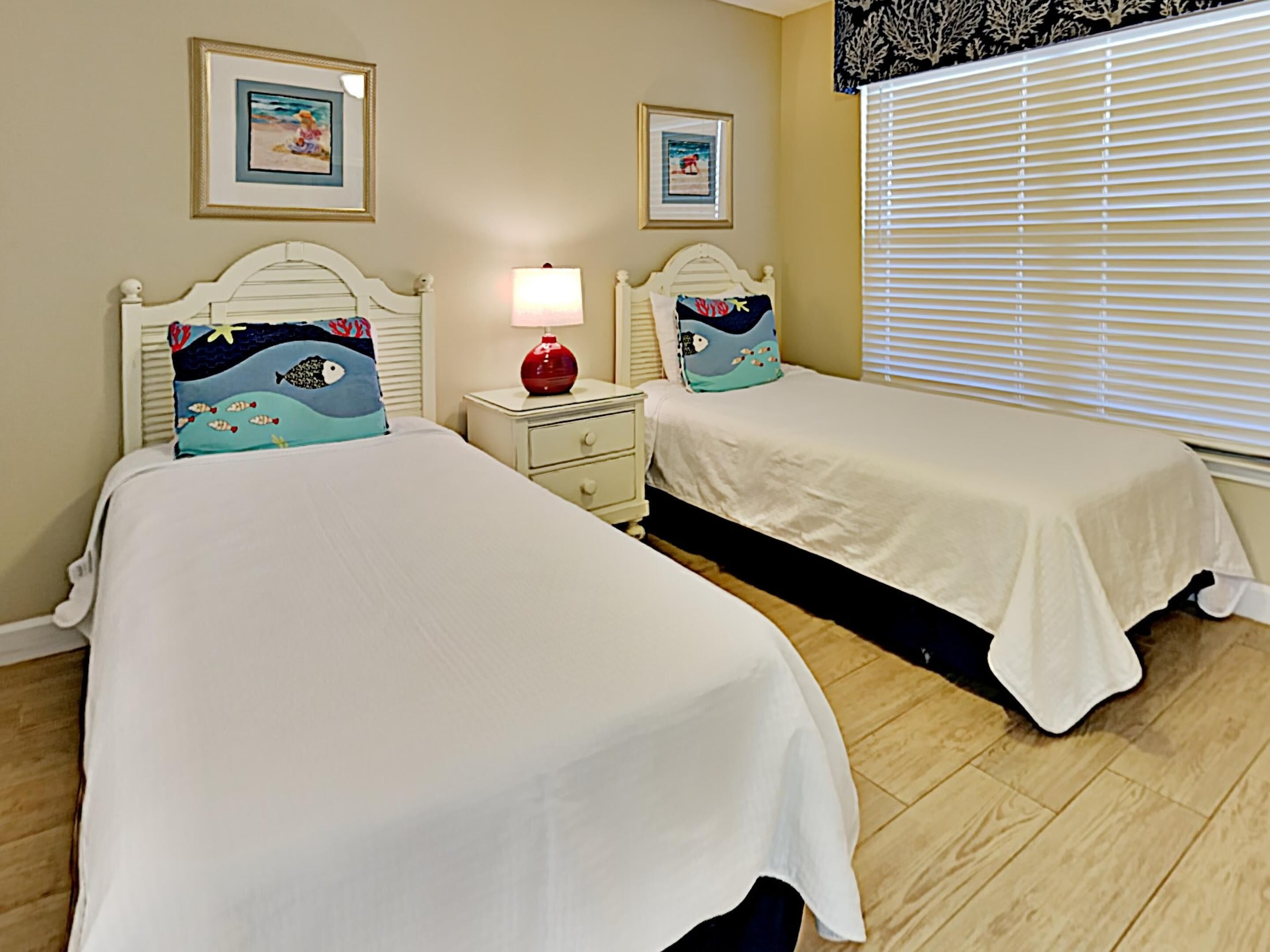 A bedroom with two beds, each with a white bedspread and matching headboards. There are two bedside tables with lamps and decorative pillows. The room has wooden flooring and a window with blinds.