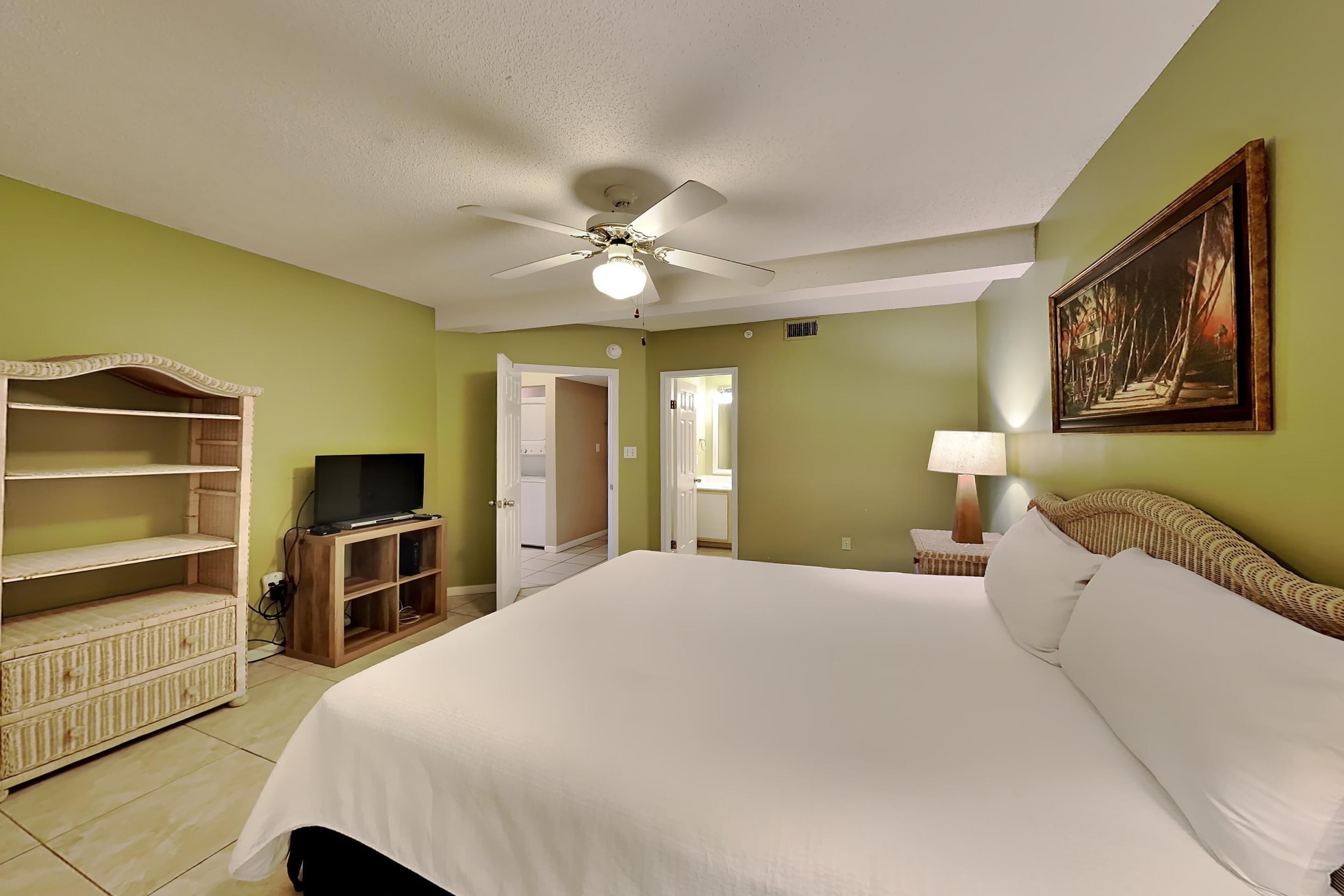 Habitación de hotel con una cama grande, ventilador de techo, televisor sobre un soporte de madera y un cuadro en la pared.