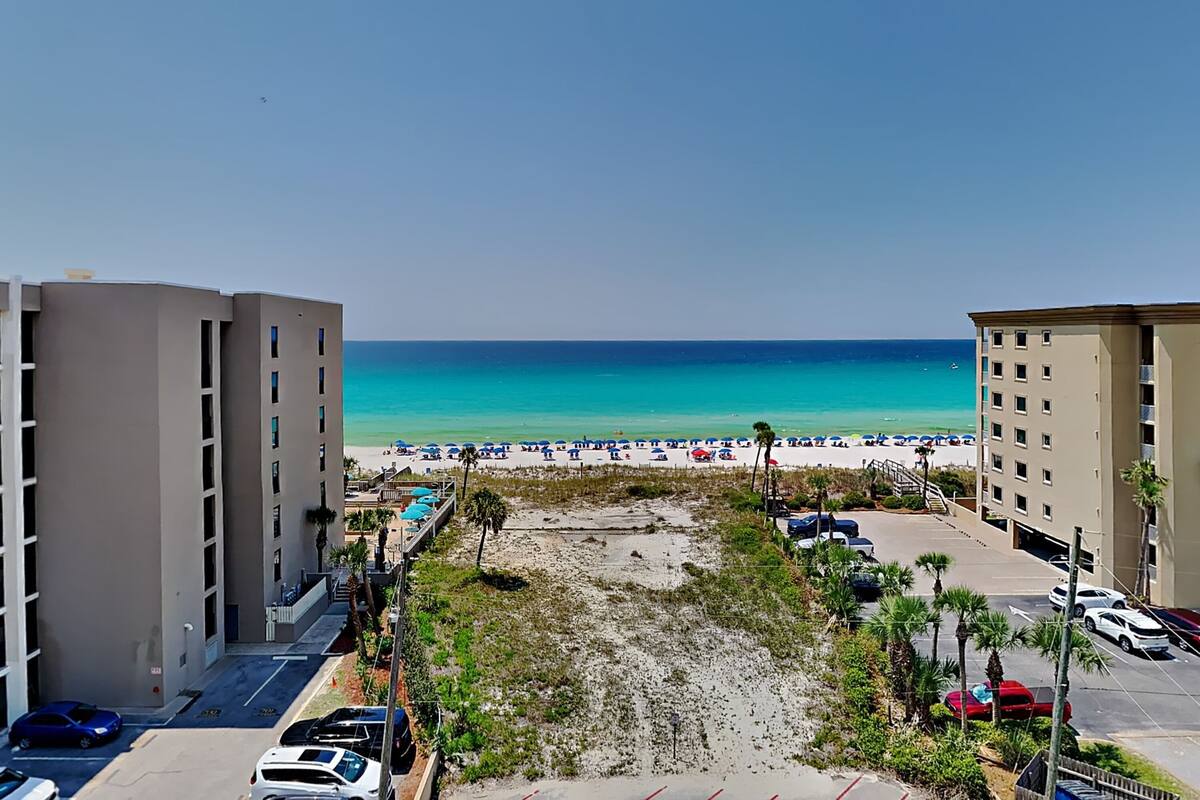 A beachfront hotel with multiple floors, a parking area, and a beach with umbrellas.