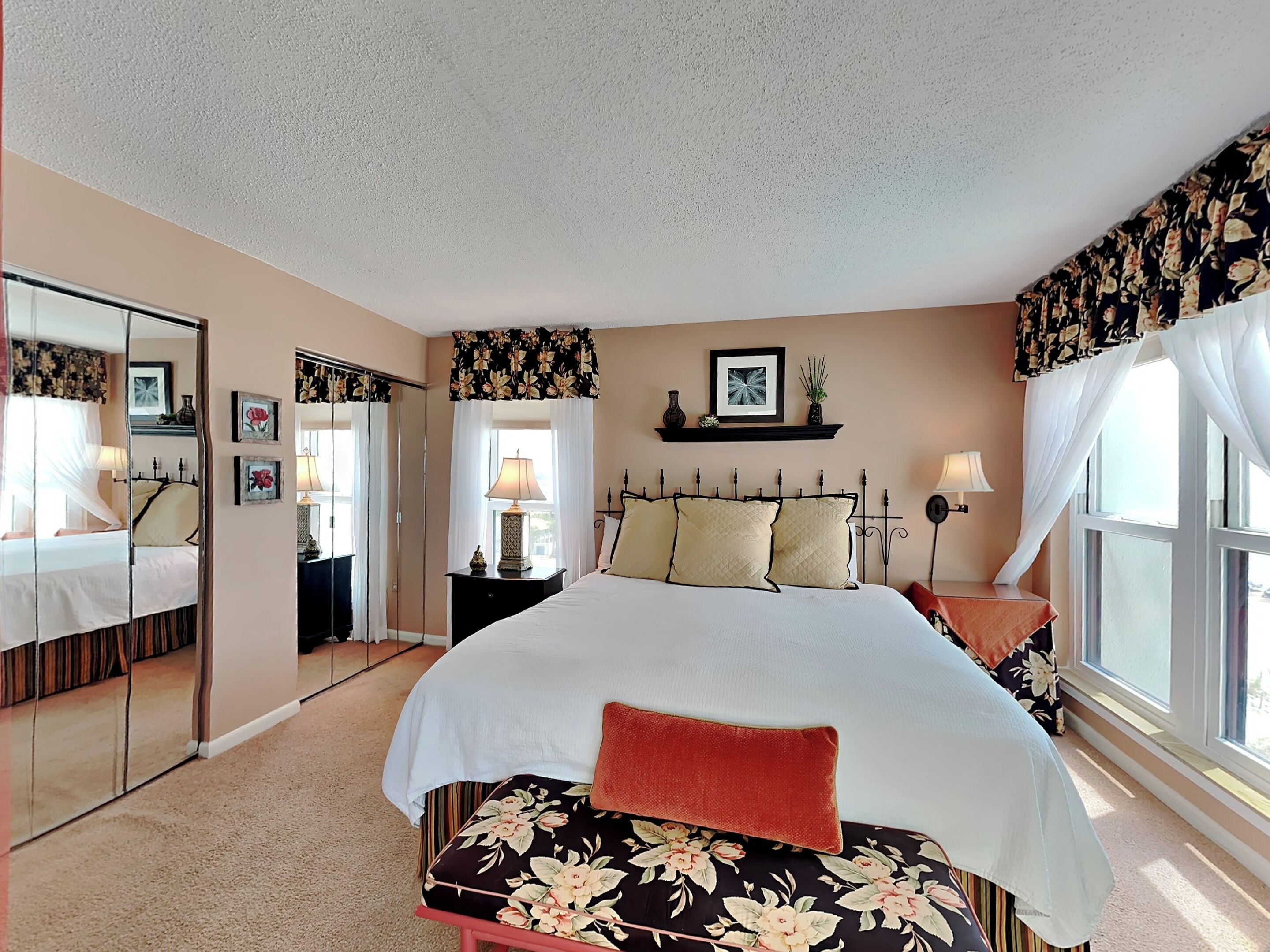 A bedroom with a bed, a nightstand, a mirror, a window with curtains, and a wall-mounted shelf with a picture.