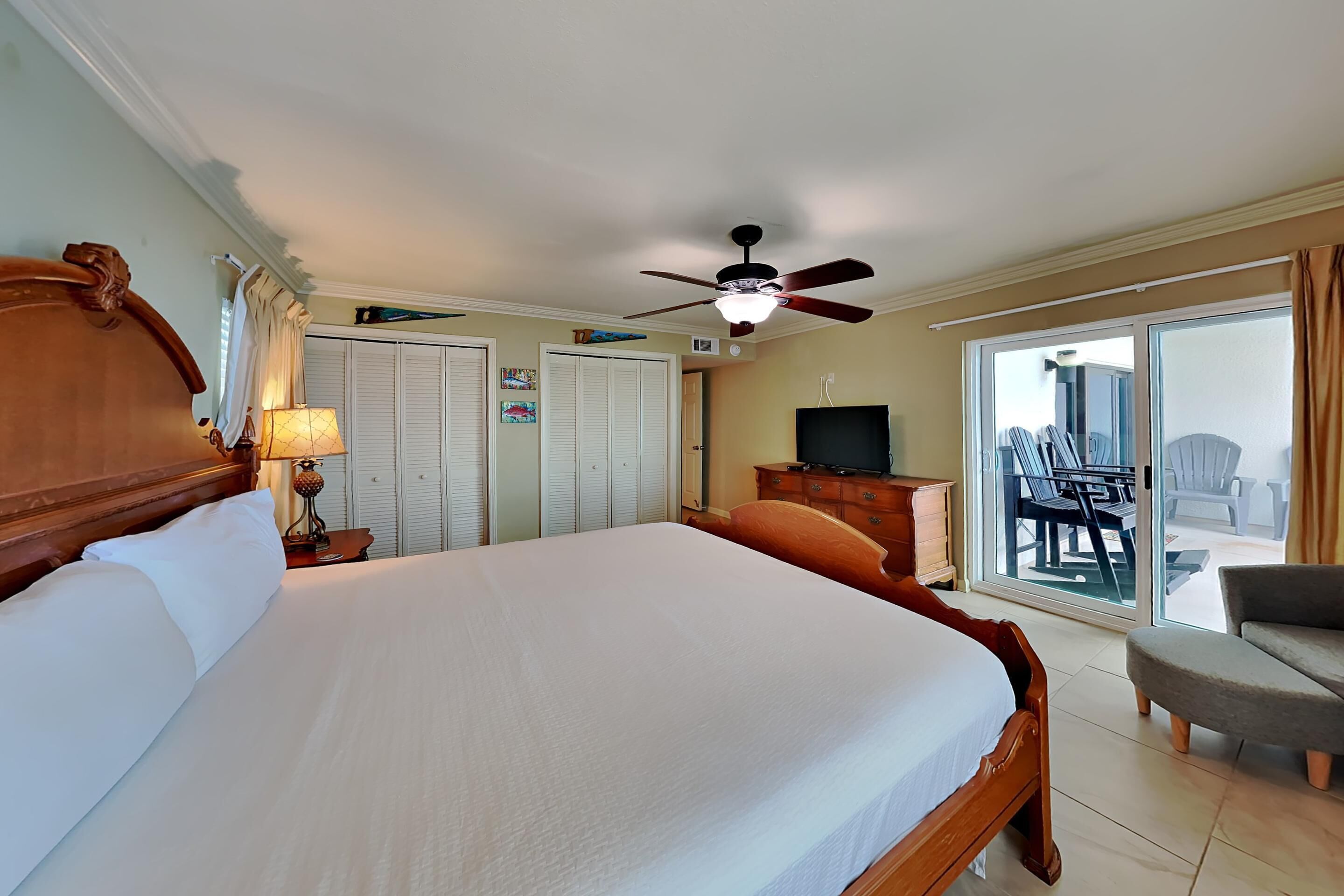 A hotel room with a large bed, a ceiling fan, a TV, a balcony with chairs, and a sliding glass door.