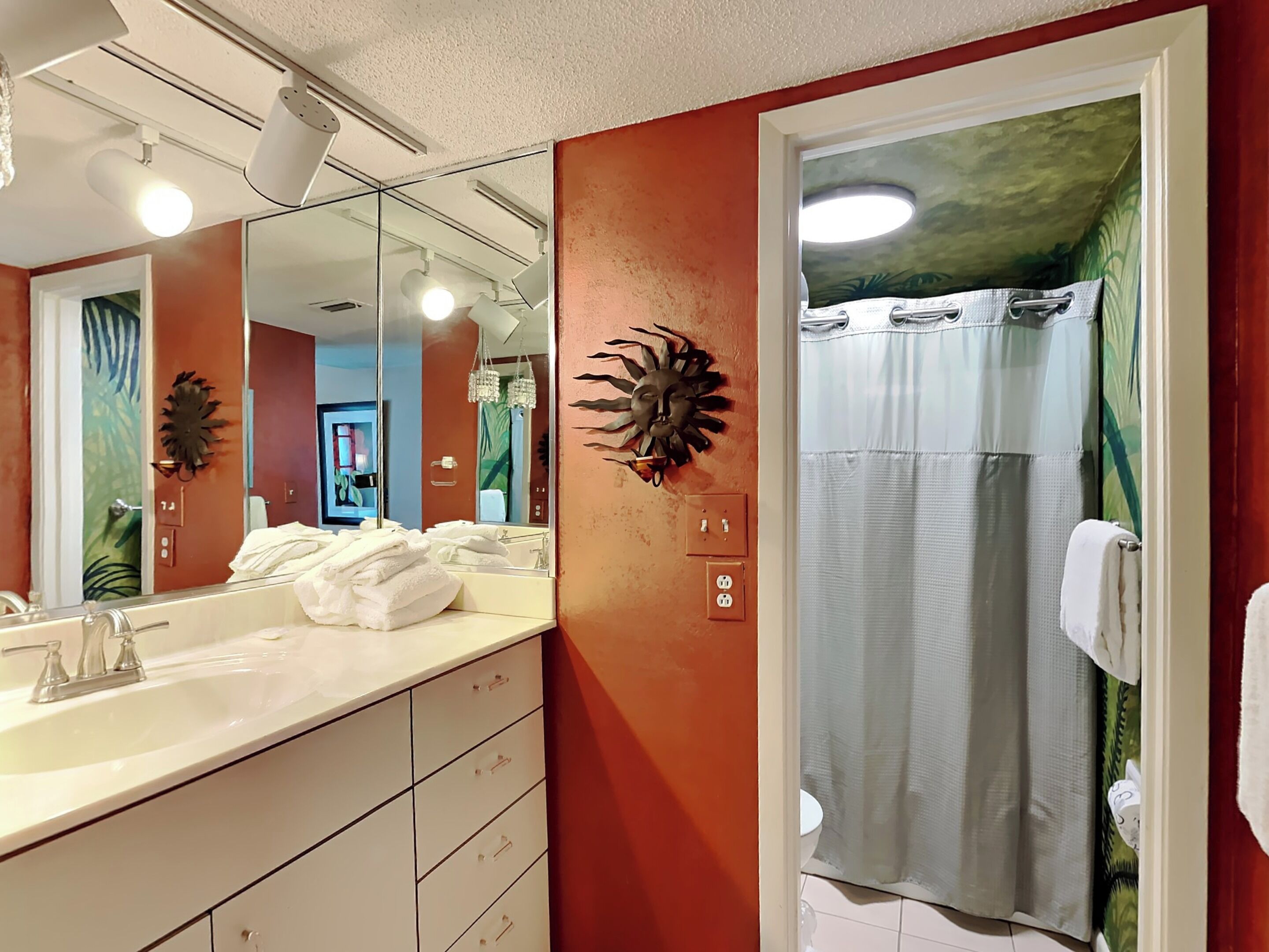 A bathroom with a white sink, mirror, and towel rack. There is a shower curtain with a tropical mural, and a wall-mounted light fixture.