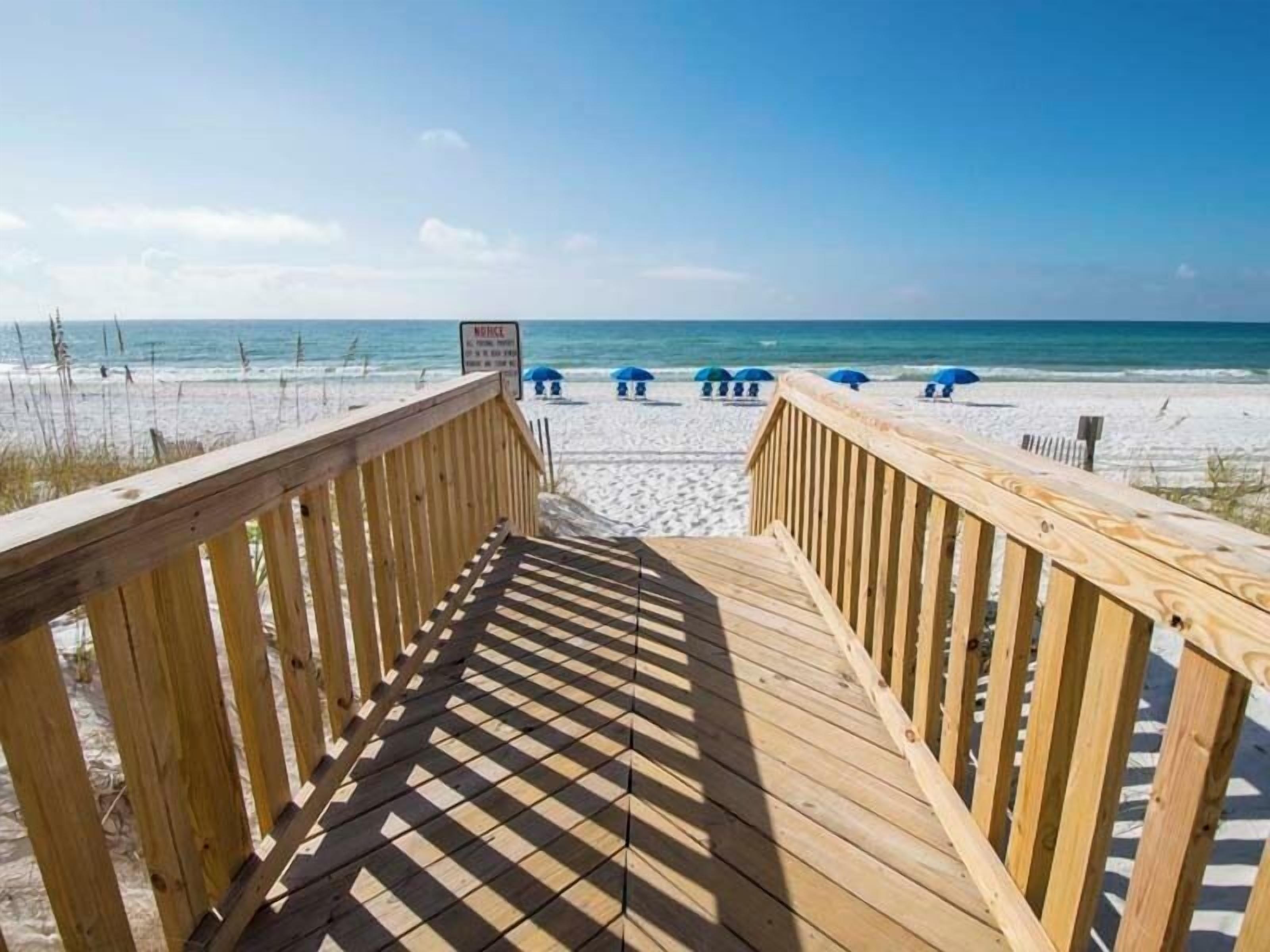 Pasarela de madera que conduce a una playa con sombrillas y un letrero que indica 'No nadar'.
