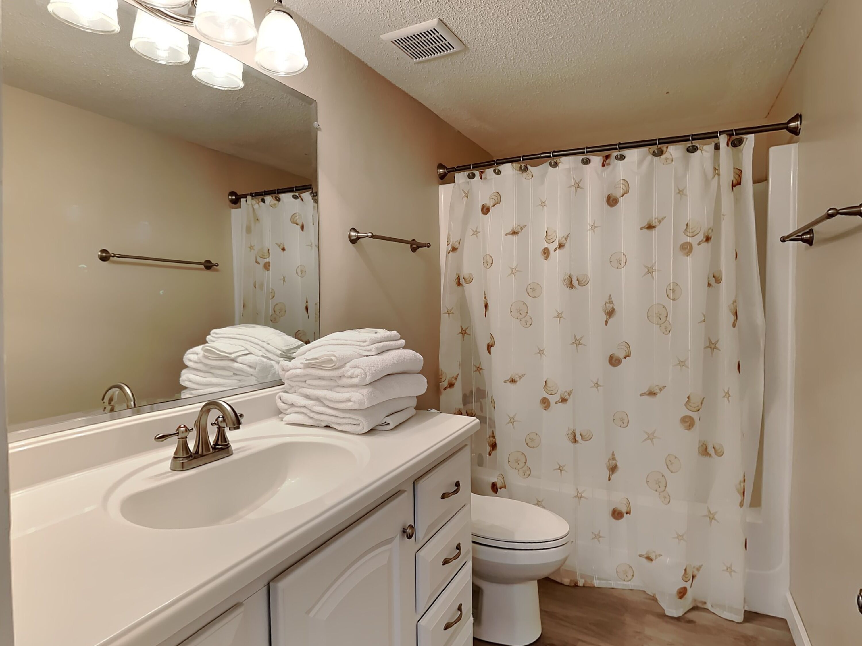 Une salle de bain équipée d’un lavabo, d’un miroir, d’une pile de serviettes, de toilettes et d’un rideau de douche orné d’un motif de coquillages.