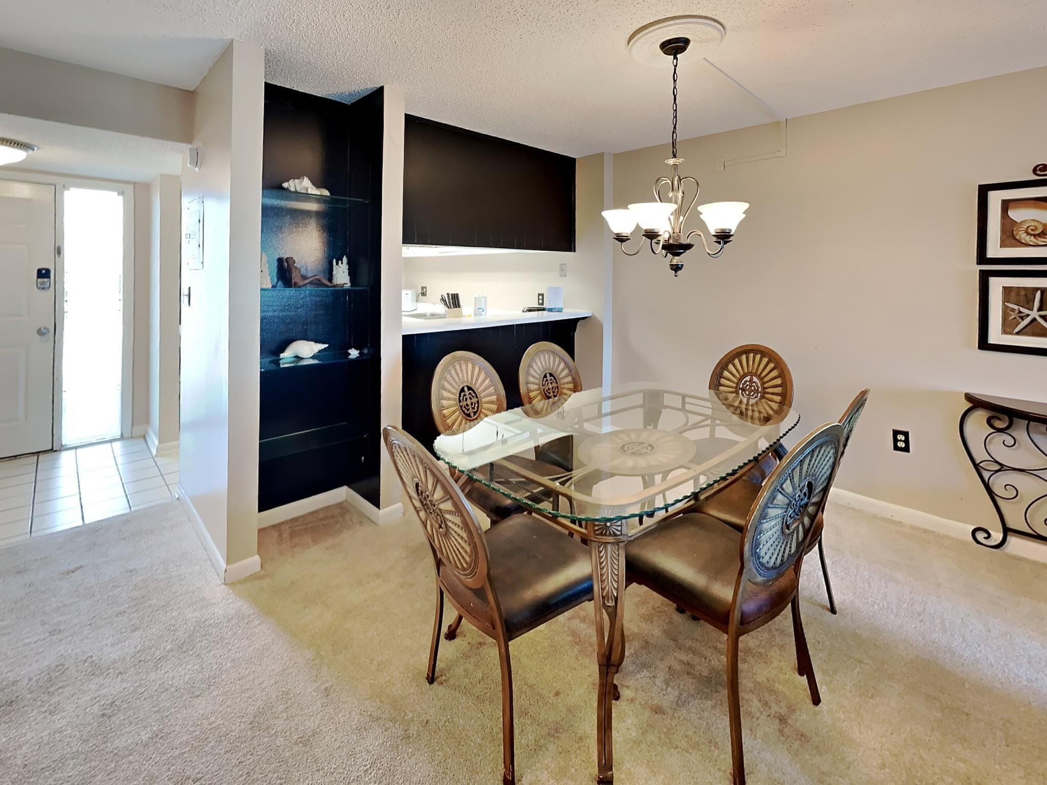 A dining area with a glass table and wooden chairs, a chandelier, and a black shelving unit with decorative items.
