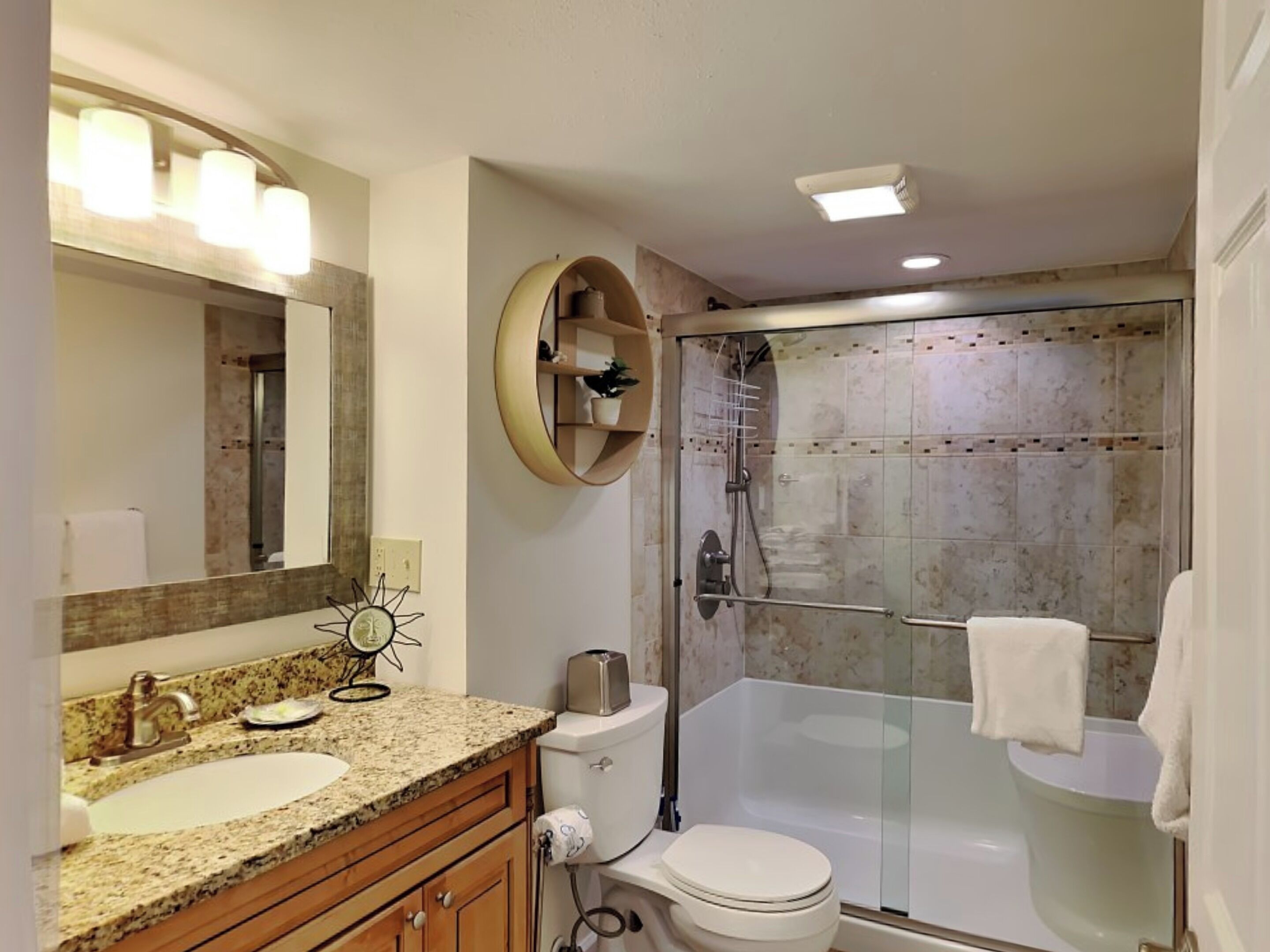 Un baño con lavamanos de encimera de granito, inodoro, ducha con mampara de vidrio y espejo con adorno en forma de sol.