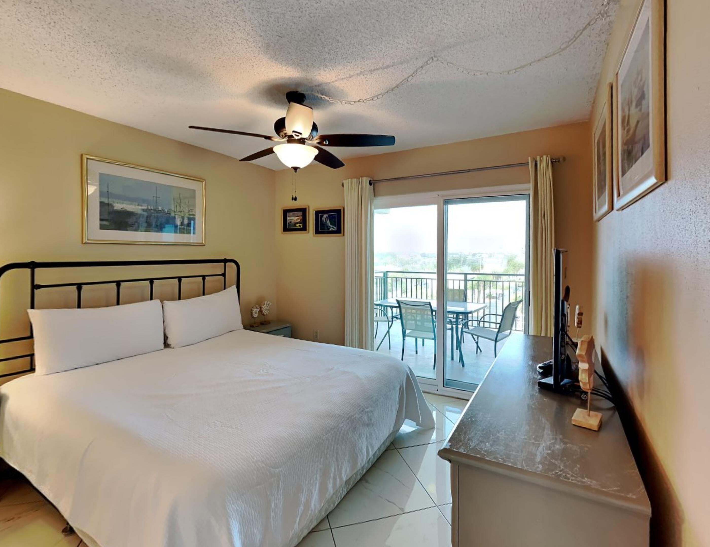 A hotel room with a bed, a ceiling fan, a balcony with chairs, and a view of the outdoors.