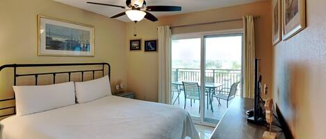 A hotel room with a bed, a ceiling fan, a balcony with chairs, and a view of the outdoors.