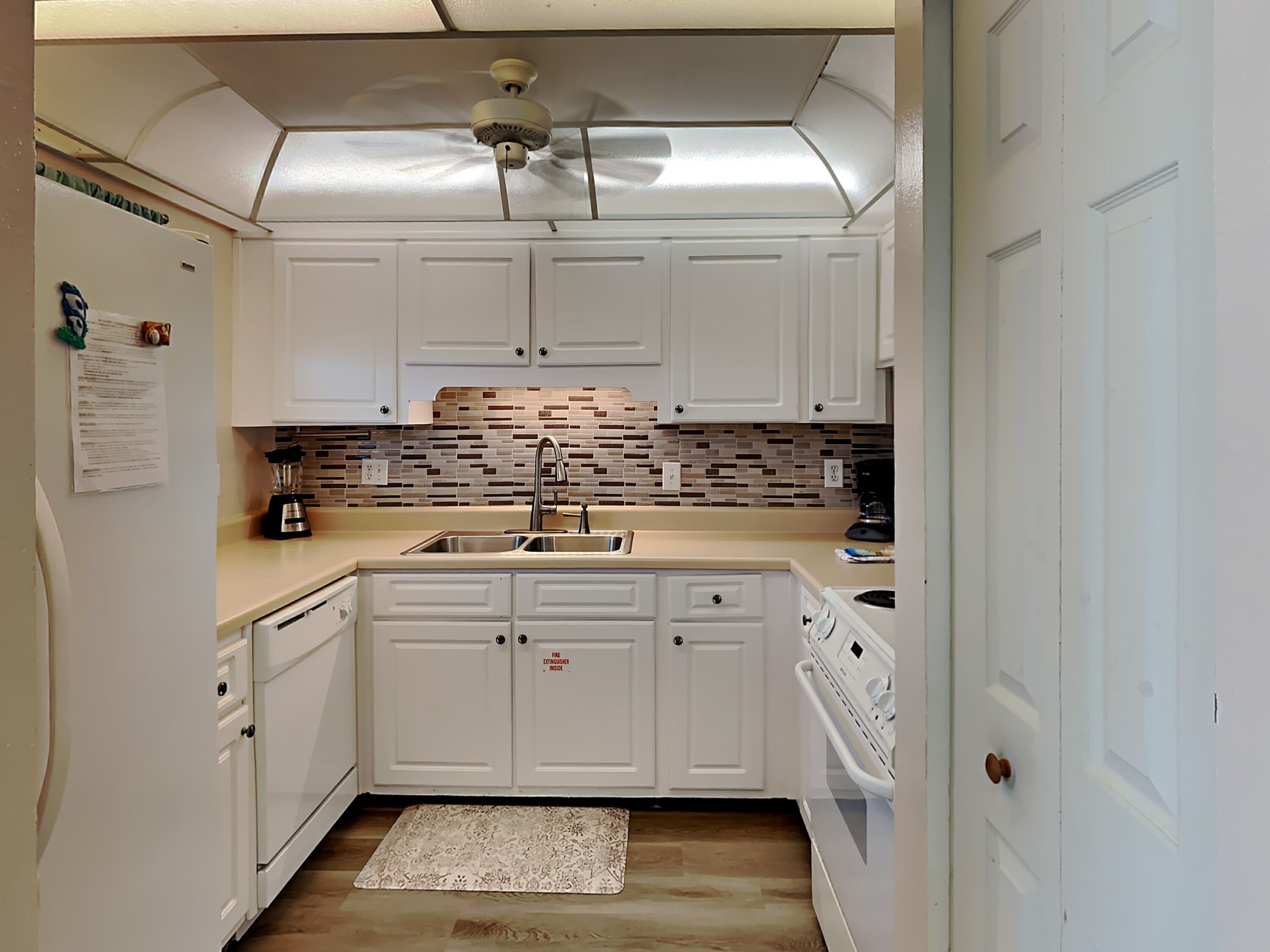 Una cocina compacta con armarios blancos, salpicadero de azulejos, horno empotrado y ventilador de techo.