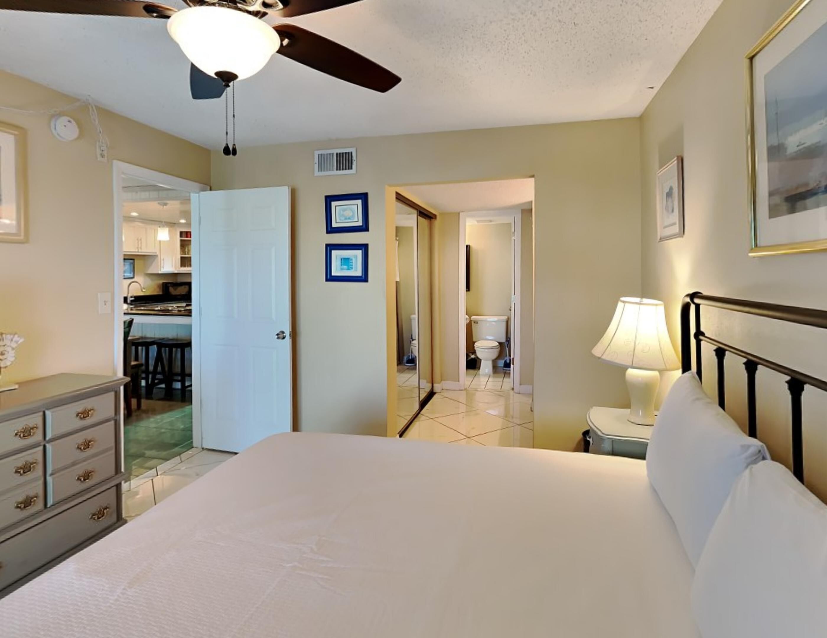 A bedroom with a bed, a nightstand, a ceiling fan, and a view of the kitchen and bathroom through an open door.