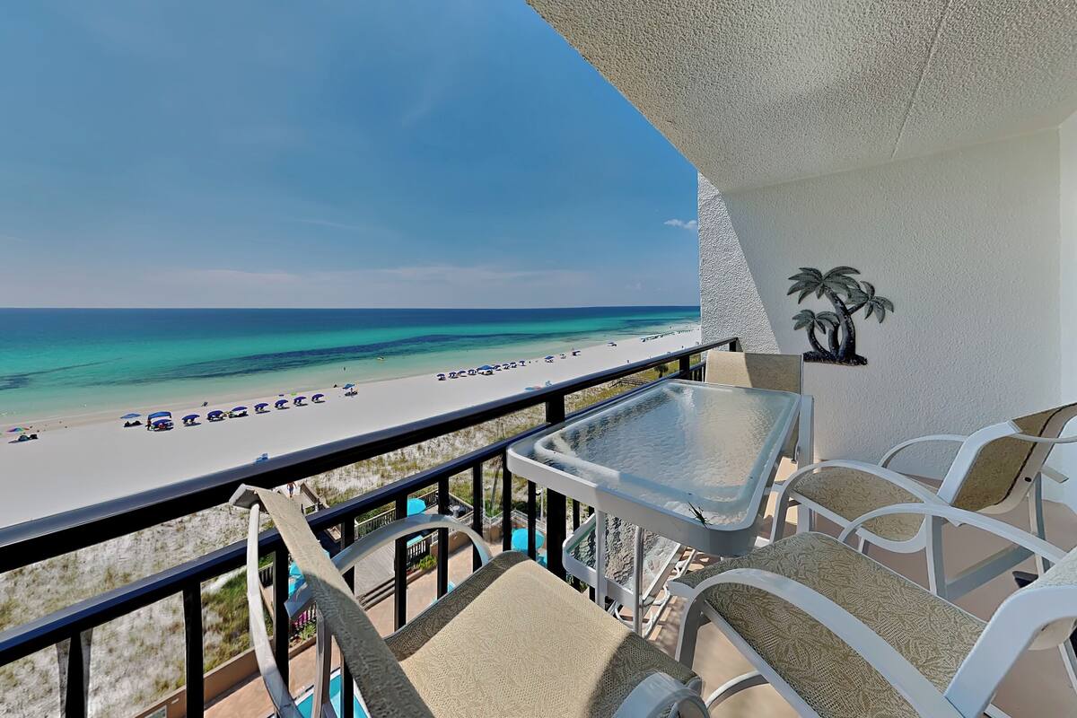 A balcony with a view of a beach, white outdoor furniture, and a palm tree decoration.