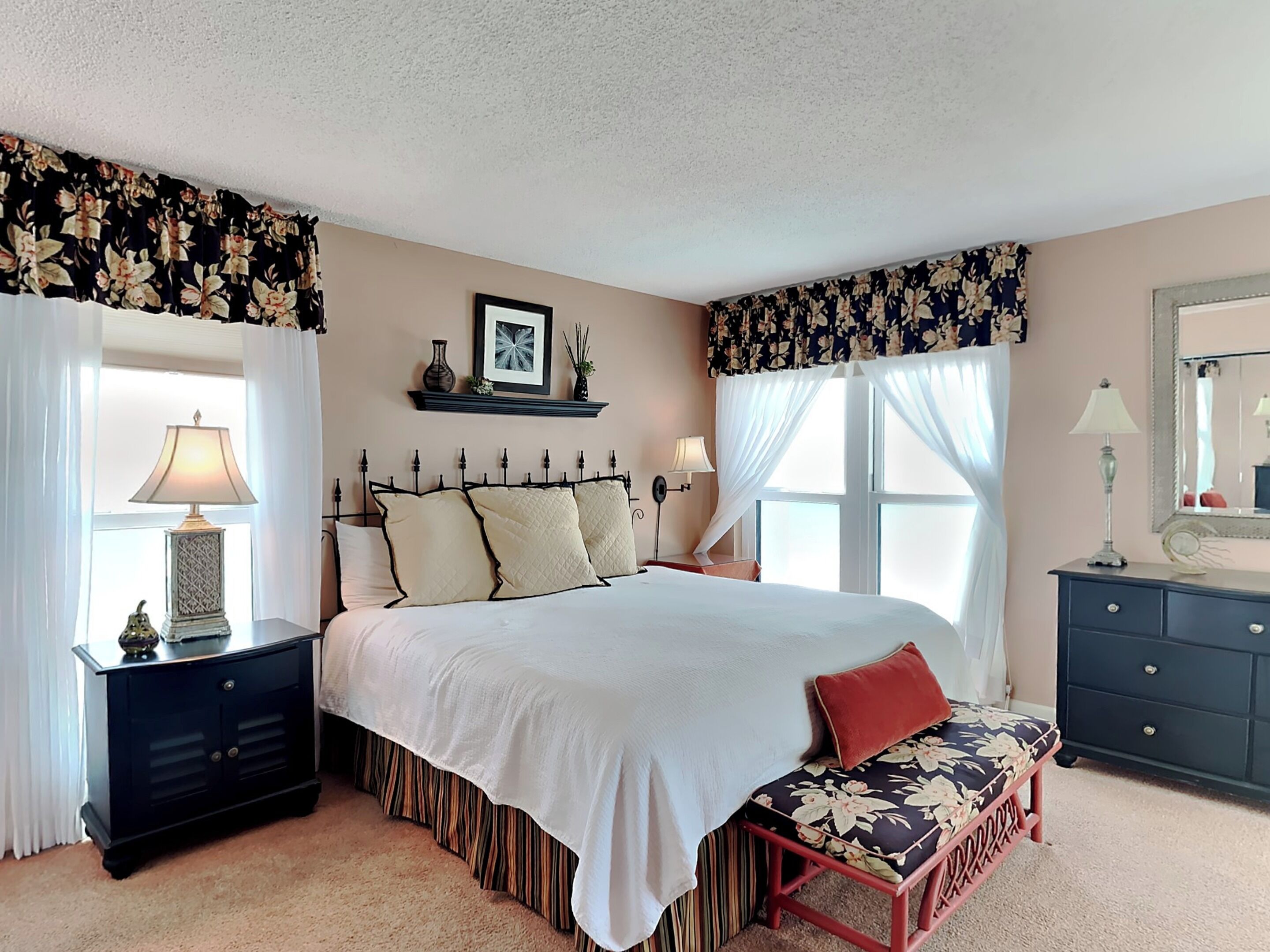 A bedroom with a large bed, two bedside tables, a bench, a dresser, and a window with curtains.