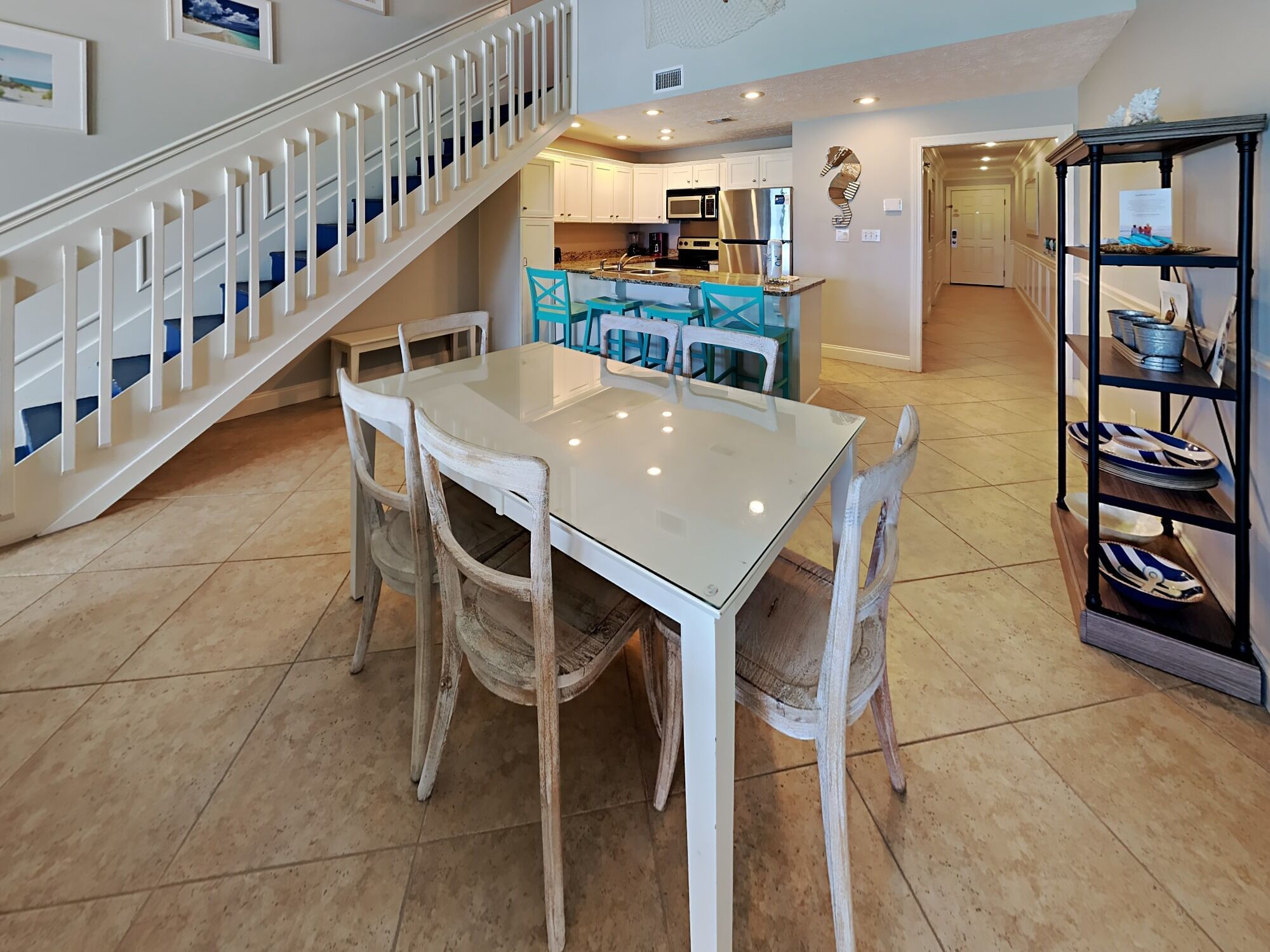 A dining area with a white table and chairs, a kitchen in the background, and a staircase with white railings.