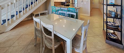 A dining area with a white table and chairs, a kitchen in the background, and a staircase with white railings.