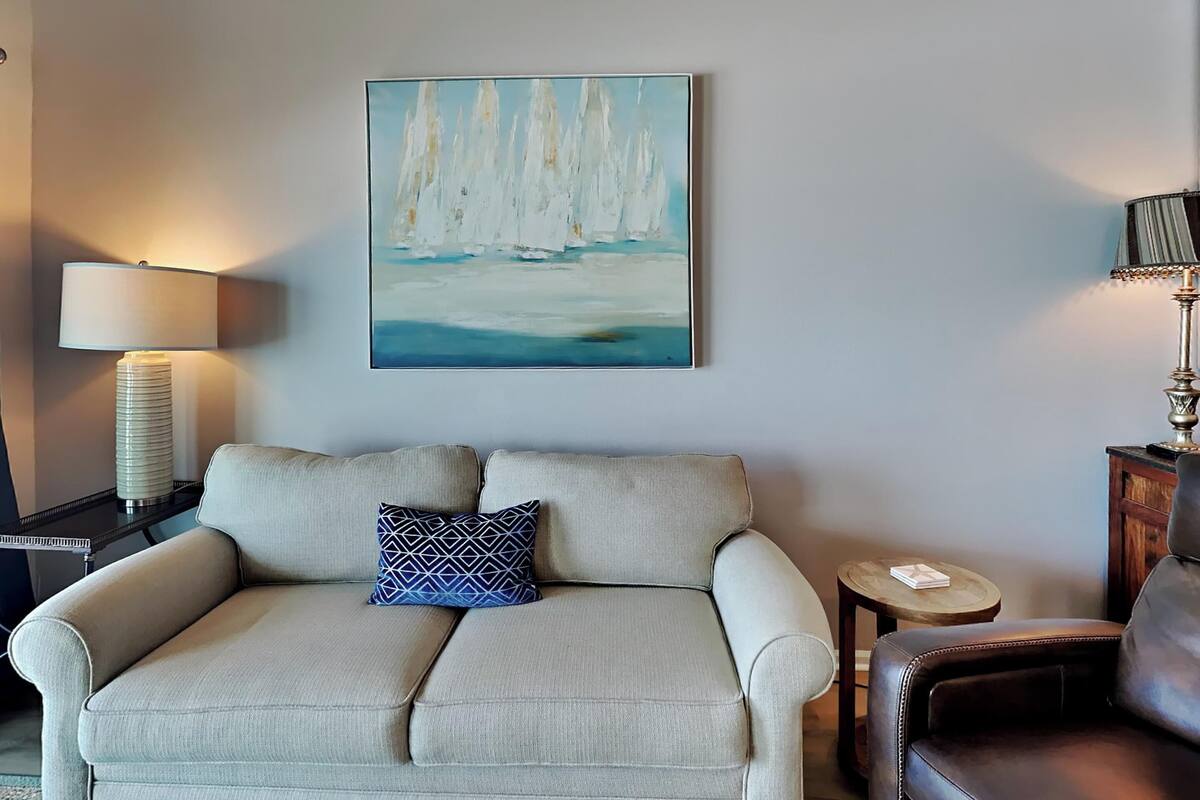 Un salon avec un canapé beige, un coussin à motifs, un tableau représentant des voiliers, une lampe et une petite table.