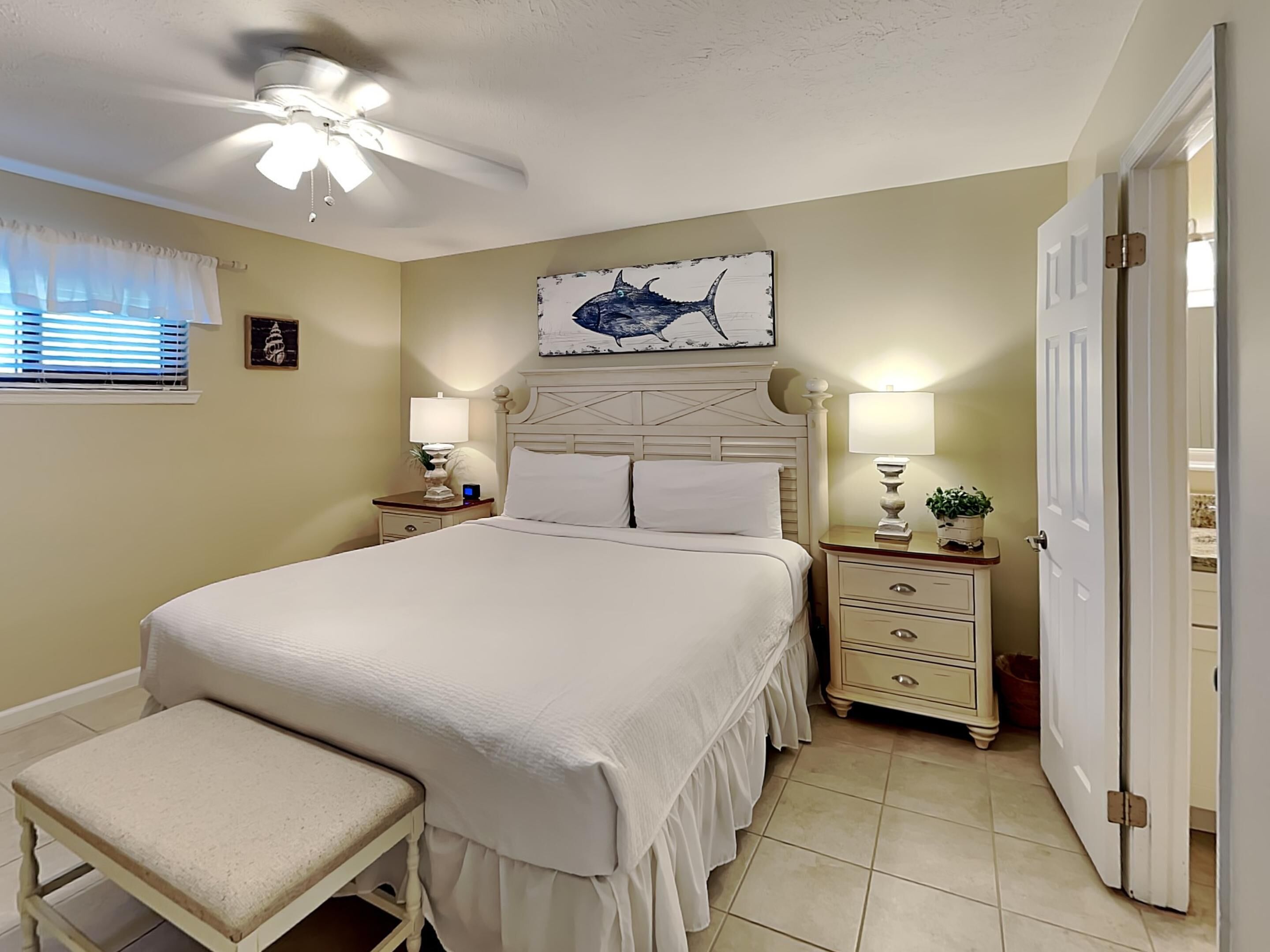 A bedroom with a bed, a bench, a ceiling fan, a wall-mounted artwork, and a door.
