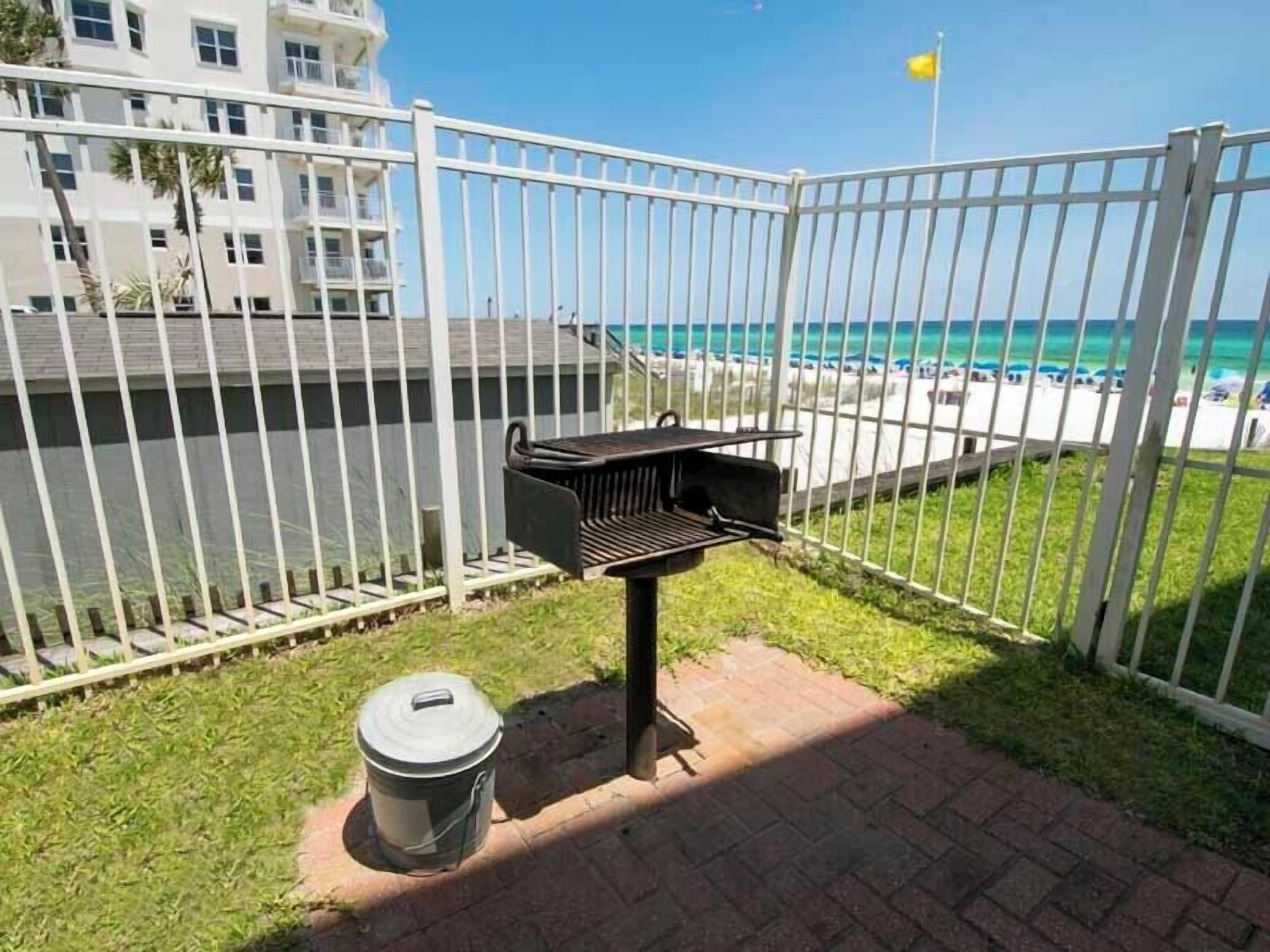 A barbecue grill and trash can in a fenced outdoor area with a view of the beach and ocean.