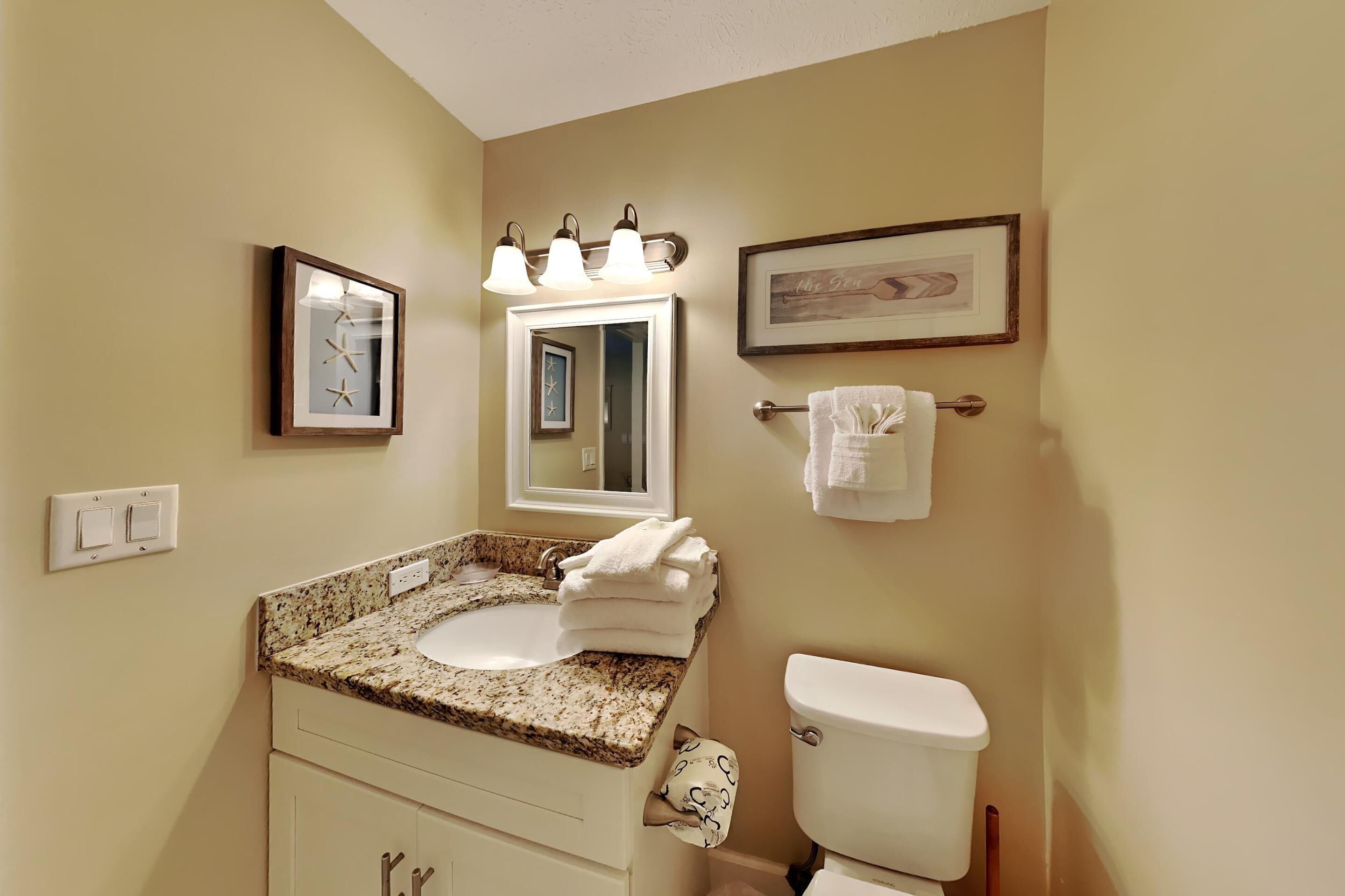 Une salle de bain avec un lavabo, un miroir et des toilettes. Des tableaux encadrés ornent le mur, ainsi qu’un porte-serviettes et un luminaire placé au-dessus du miroir.