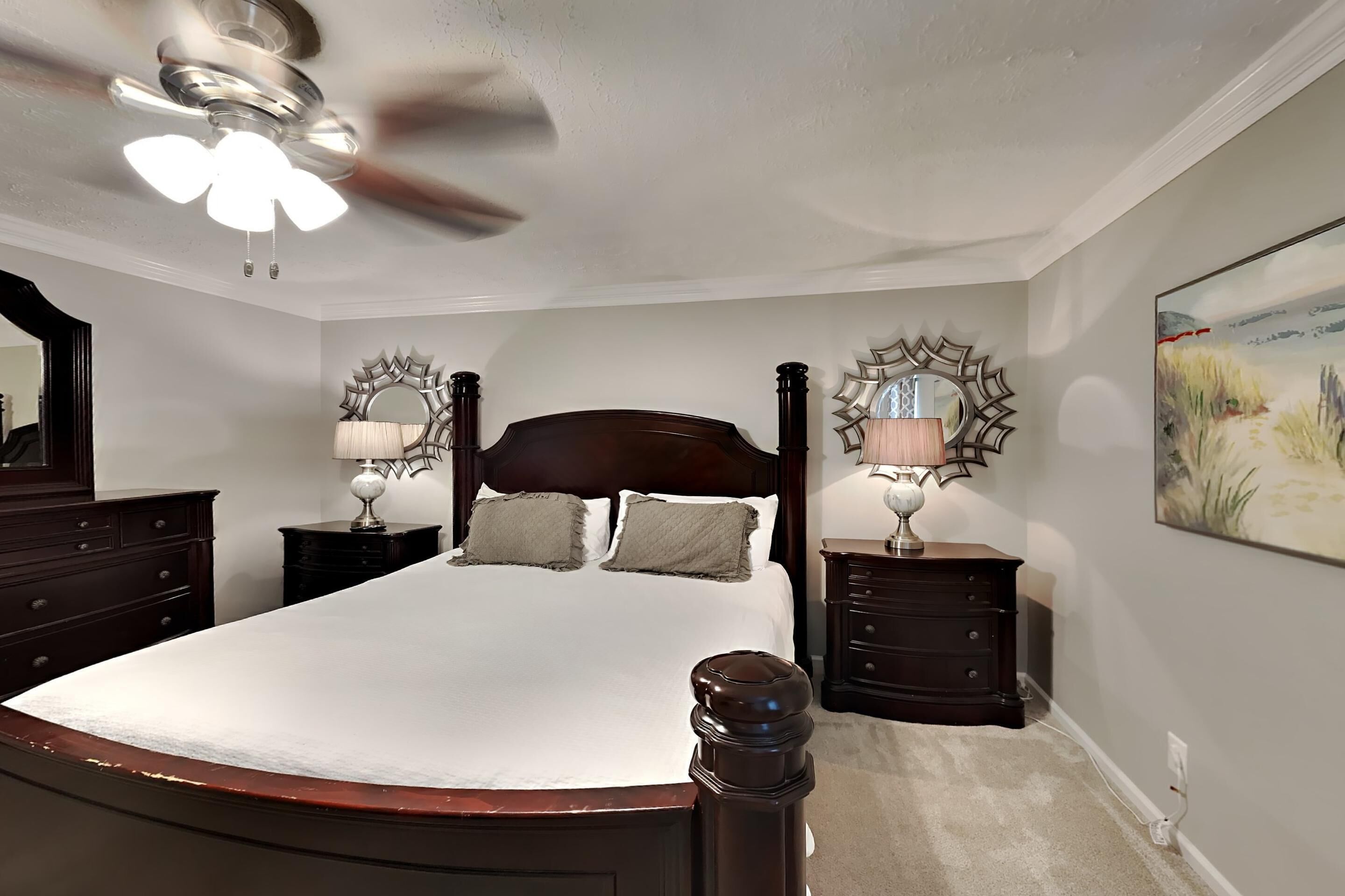 Une chambre à coucher avec un grand lit, deux tables de chevet avec des lampes, un ventilateur de plafond et un tableau au mur.