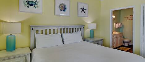 Un dormitorio con una cama, dos mesitas de noche con lámparas, un tocador, un ventilador de techo y un baño visible a través de una puerta abierta.