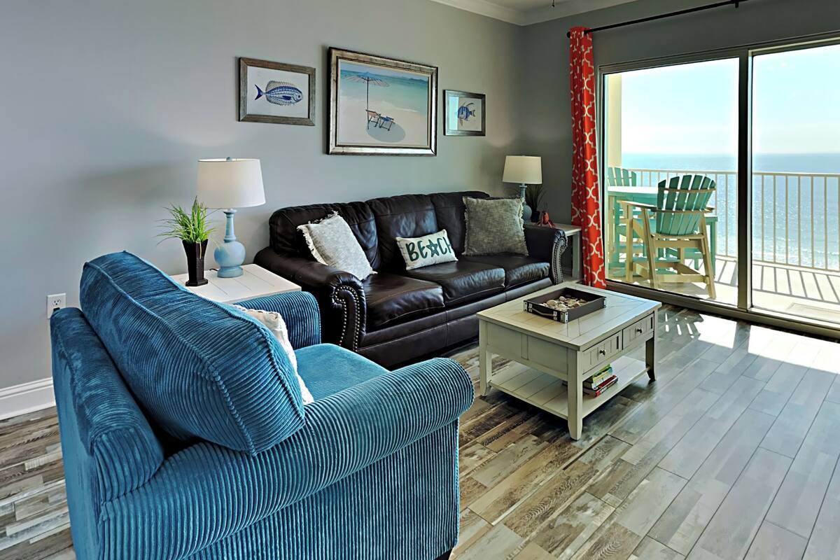 A living room with a black leather sofa, a blue sofa, a coffee table, and a view of the ocean through large windows.