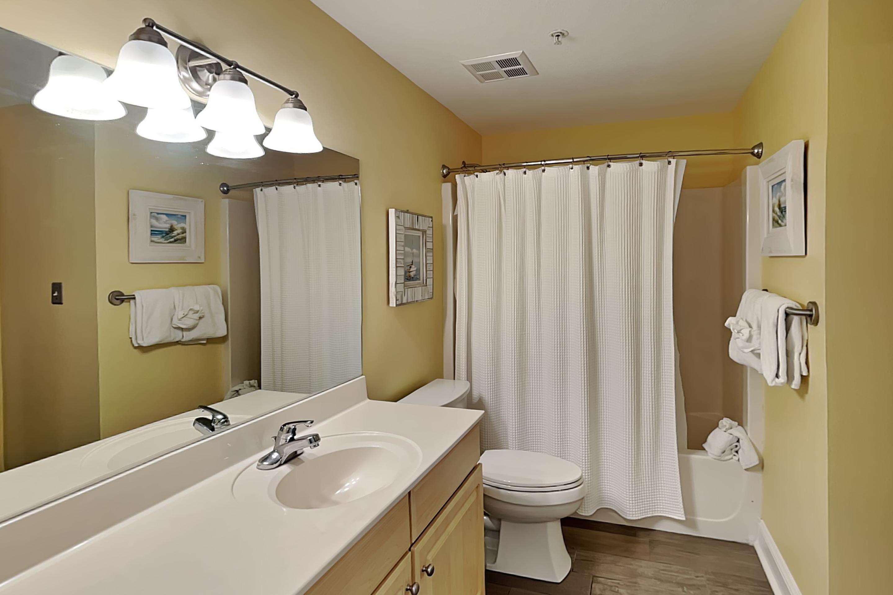 A bathroom with a large mirror, a sink, a toilet, a shower curtain, and towels on the wall.