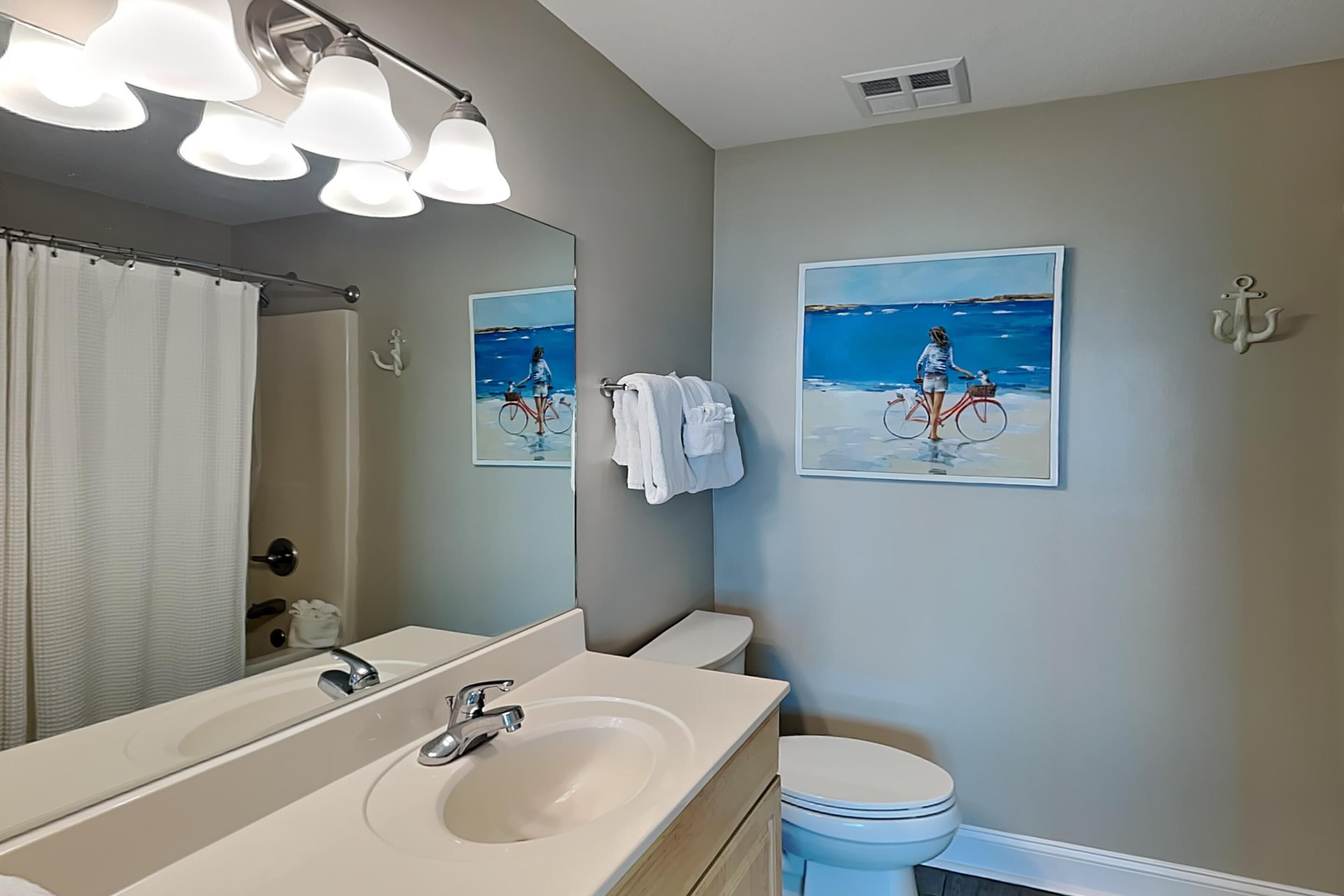 Une salle de bain avec un grand miroir, un lavabo et des toilettes. Deux tableaux encadrés sont accrochés au mur et représentent une scène de plage avec une personne à vélo.
